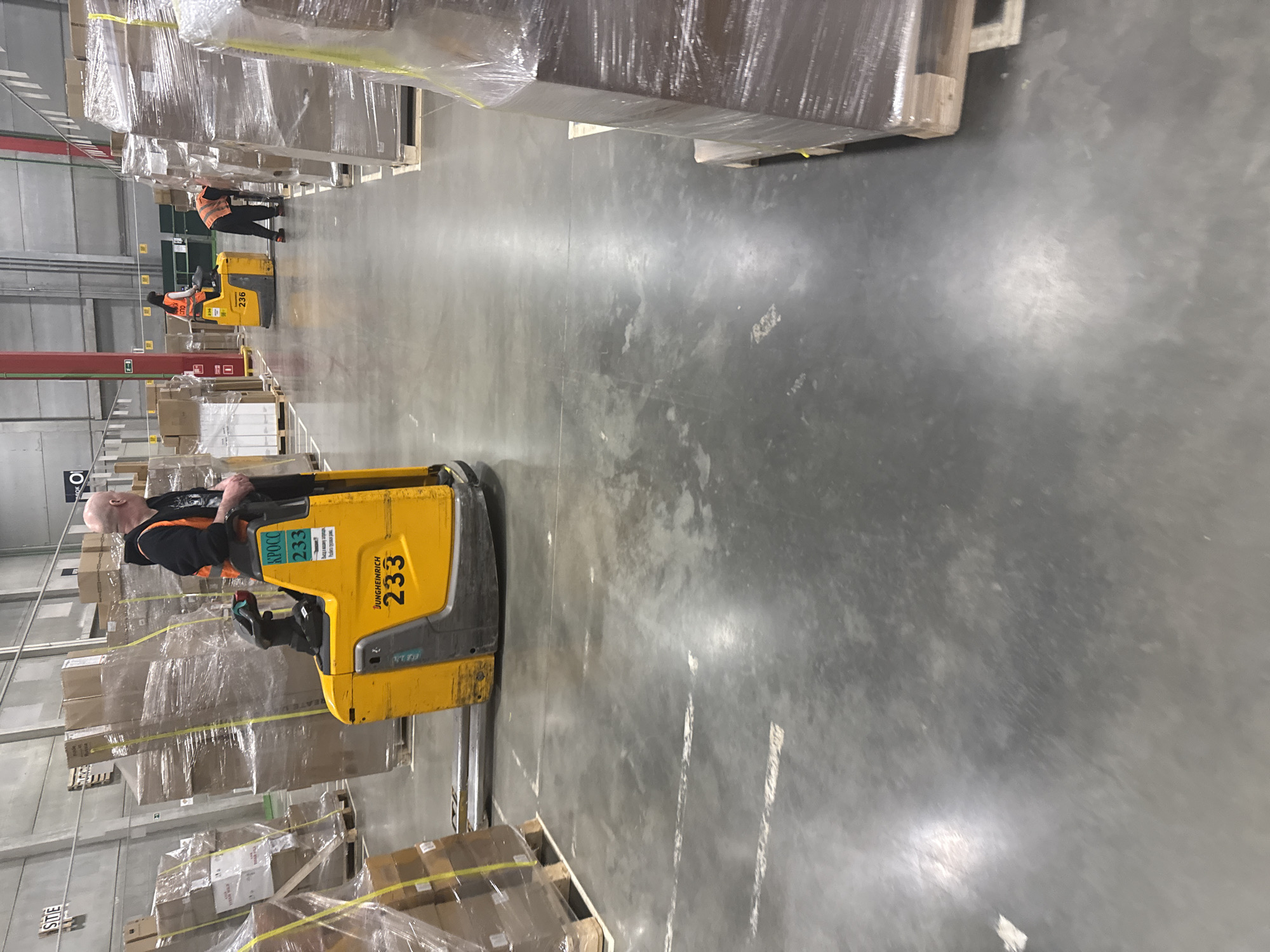 The image depicts the interior of a large warehouse in Lupanovo, Russia. The warehouse features a concrete floor and tall walls lined with shelves stacked with boxes and goods wrapped in plastic. The scene includes one worker driving a yellow Jungheinrich forklift, number 233, loaded with shrink-wrapped packages. In the distance, another forklift and its operator are visible, also transporting materials. The overall setting suggests a distribution or storage facility within an industrial area.