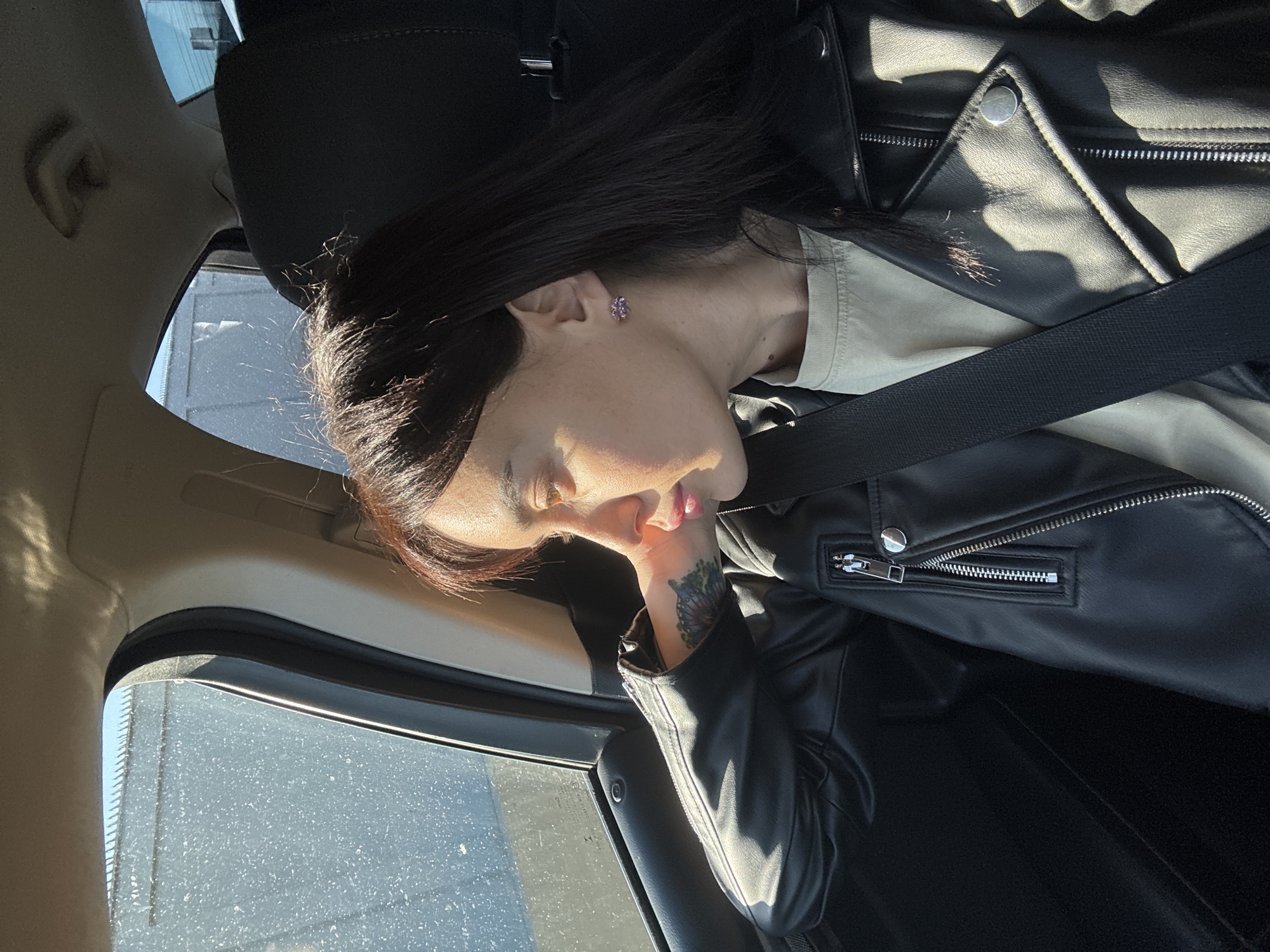 A woman rests her head against the window of a car. She has long, dark hair, fair skin, and a small purple earring. She is wearing a black leather jacket and a light-colored shirt. A black seatbelt is positioned diagonally across her body. Her hand, adorned with a floral tattoo, rests near her face. The car window is partially covered with grime or dust. The background suggests an interior car setting during daylight. There is insufficient detail to determine the location in Tbilisi, Georgia.