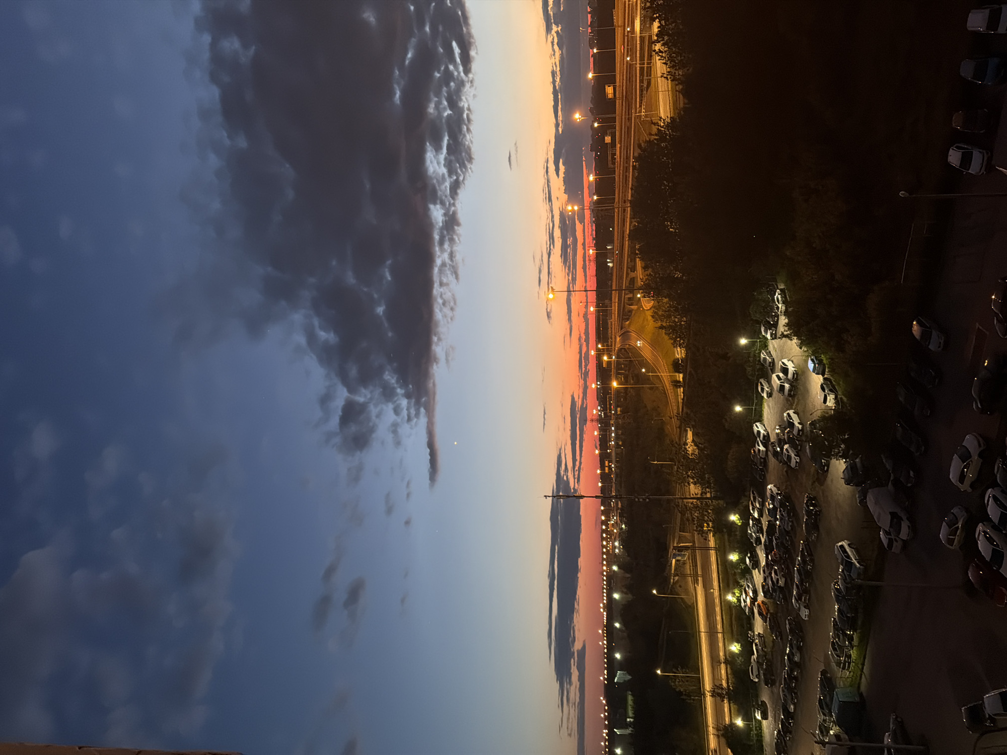 This is a high-angle, wide shot of a city scene at dusk. The sky is a gradient of deep blue at the top, transitioning to orange and pink near the horizon, with dark clouds scattered across the upper portion. A highway with multiple lanes and streetlights is visible in the mid-ground, curving through the landscape. In the foreground and mid-ground, there are several large parking lots filled with numerous cars. The parking lots are illuminated by streetlights, casting a warm glow. Trees and other foliage are visible between the parking lots and the highway, and also in the far background, partially obscuring distant buildings. The overall scene depicts a suburban or industrial area of a city transitioning from day to night.