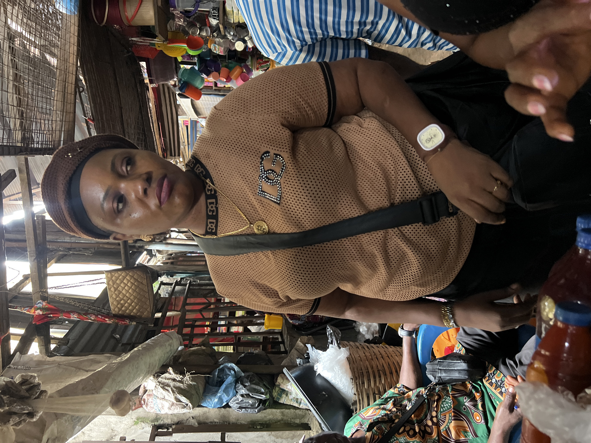 A medium shot captures a woman in a brown mesh shirt and beret in an open-air market stall in Nkpor, Nigeria. She wears a gold chain with a Bitcoin pendant and a dark crossbody bag. To her left, another person in a green and orange patterned shirt is visible. In the background, shelves are stocked with various items, including buckets, containers, and what appear to be bottles of drinks in the foreground. The market's structure, with its wooden beams and corrugated iron roof, suggests an outdoor, possibly makeshift, setting. The lighting is diffused, indicating an overcast day or a shaded area within the market.