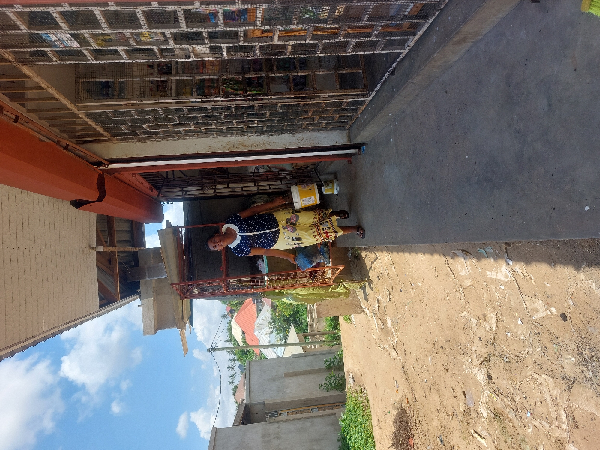 The scene captures a middle-aged woman standing outside a storefront in a sunny environment. She wears a dark blue and white polka-dot dress and a yellow skirt with a printed pattern. The woman stands behind a metal cage enclosure.
The background reveals buildings under a blue sky with scattered clouds, suggesting an urban or suburban area.
The overall setting appears to be Dar es-Salaam, Tanzania, based on the architectural style of the building, the climate, and the local vegetation.