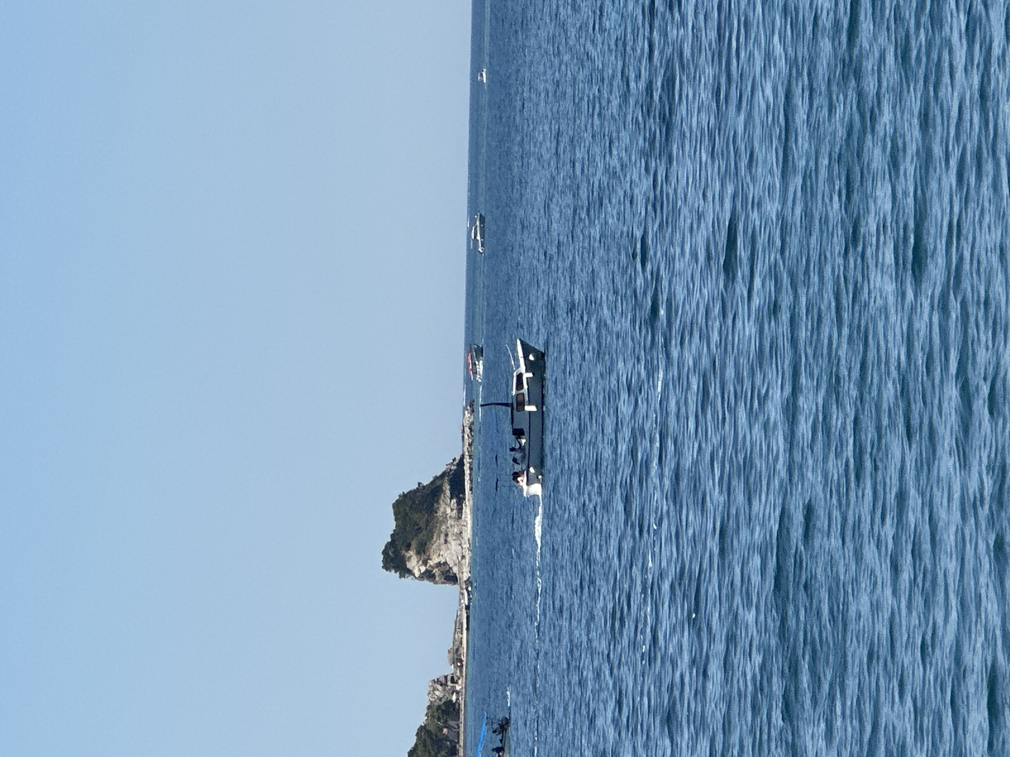 A wide, low-angle shot captures a section of the Adriatic Sea under a clear, pale blue sky. The water is a deep, rich blue, with a gentle, textured surface reflecting the bright sunlight. In the mid-ground, a white motorboat with a dark canopy is actively moving through the water, leaving a visible white wake. Several figures are seated on the boat. Further in the distance, a cluster of rocky islands covered in green vegetation rises from the sea. A few other smaller boats are scattered across the horizon, some moving and some stationary. The scene is tranquil and suggests a sunny day in a coastal area, likely near Budva, Montenegro, given the distinctive rocky islands.