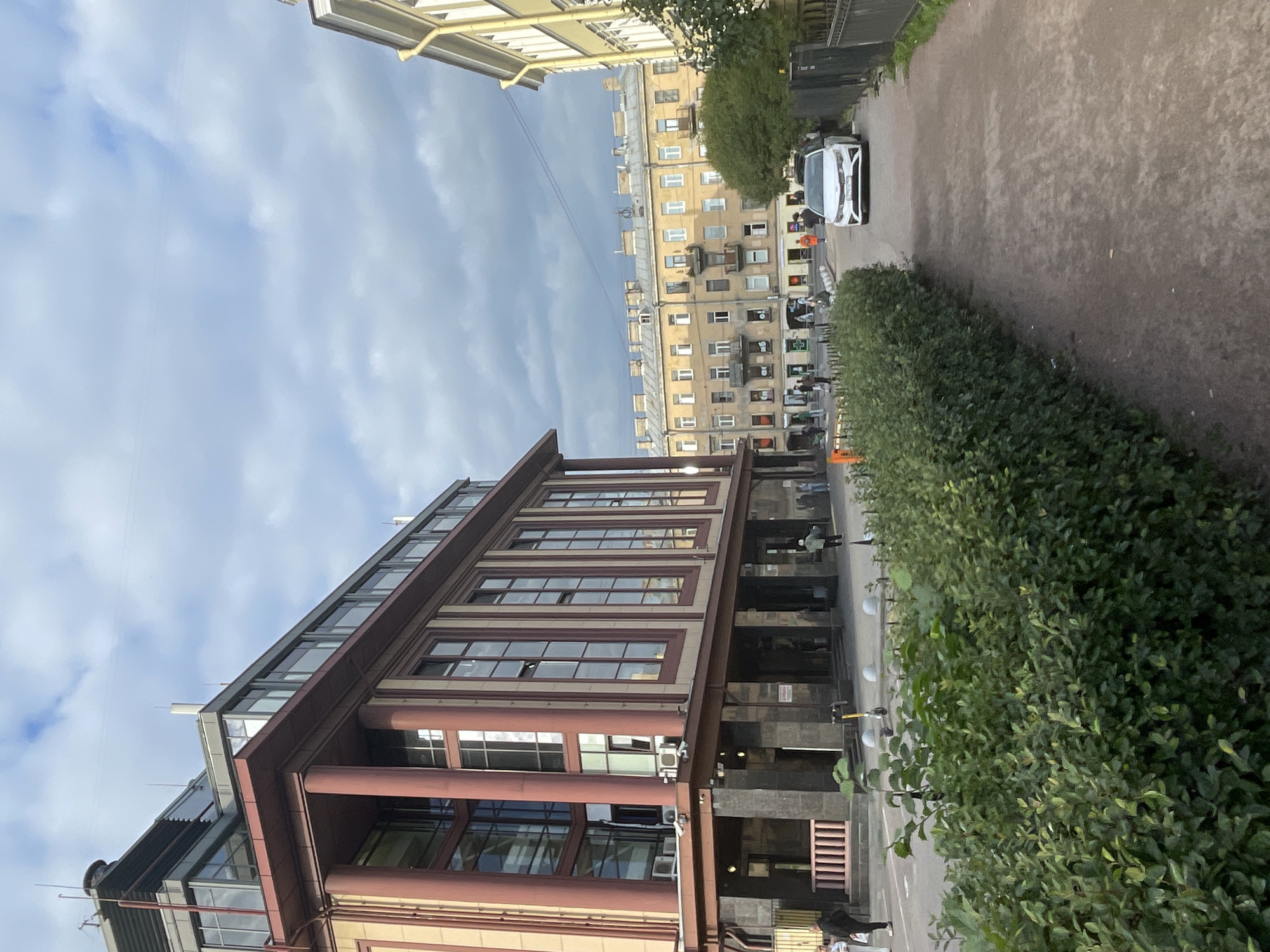 The image depicts an urban street scene in Saint Petersburg, Russia. The foreground is dominated by a dense, manicured green hedge lining a gravel path. Beyond the hedge, a light-colored modern car is parked on the street. To the left, a large, multi-story building with a red-brown facade and substantial columns occupies a significant portion of the frame. This building features numerous large windows and a more modern glass-enclosed section on its upper floors. Further down the street, a row of older, light-colored apartment buildings with a classical architectural style are visible, characterized by multiple windows and balconies. The sky above is partly cloudy with dramatic grey clouds mixed with patches of blue sky. Several people are visible on the sidewalk in front of the older apartment buildings, appearing to walk or stand. One person is walking towards the viewer on the left side of the frame in front of the modern building. Another person in a white jacket is standing near the white car. A yellow electric scooter is parked on the sidewalk in front of the modern building. The overall impression is that of a city street on an overcast day.
