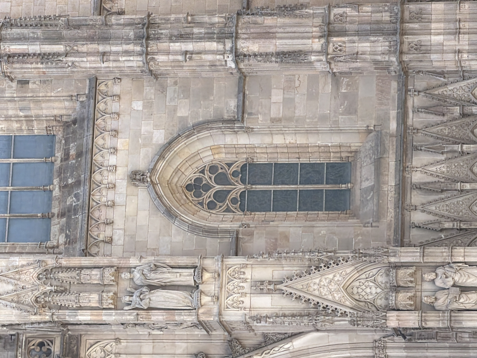 The image shows a detailed view of the exterior of a Gothic-style cathedral in Barcelona, Spain, likely the Barcelona Cathedral due to its architectural style and location. The facade features intricate stonework, including statues of figures in niches, decorative arches, and a prominent pointed-arch window with tracery. The stonework is weathered and appears to be made of limestone or a similar light-colored stone. The overall impression is one of grandeur and historical significance. The lighting suggests it is daytime, with shadows cast by the architectural features.
