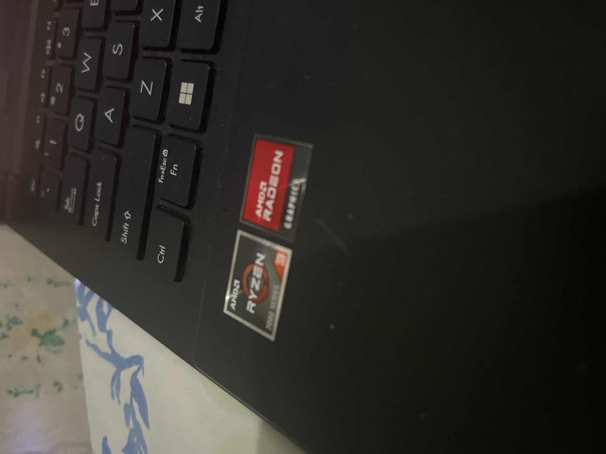 The image shows a close-up view of a laptop keyboard and the palm rest area. Two stickers are visible on the palm rest. The left sticker is square with rounded corners and features the AMD Ryzen logo with the text "7000 SERIES" below it. The right sticker is rectangular and displays the AMD Radeon logo with the word "GRAPHICS" underneath. The keyboard is black with white lettering on the keys. Some keys visible include "Caps Lock," "Shift," "Ctrl," "Fn," "Esc," "Z," "X," "Alt," and the Windows key. The background is out of focus, showing a white fabric with a blue floral pattern. There are no people or any indication of the specific location being Santa Ana, Venezuela.