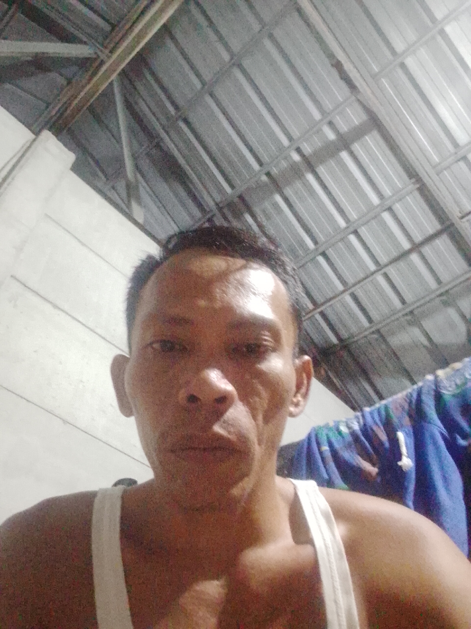 The image is a selfie of a middle-aged Asian man wearing a white tank top. He has short black hair and is gazing directly into the camera. The background includes a white wall, a metal roof structure, and a blurred blue cloth or garment hanging on a line. The location is implied to be Bukit Ketok, Indonesia, based on the user request. The man is in an interior space. The details are limited due to image quality and composition.