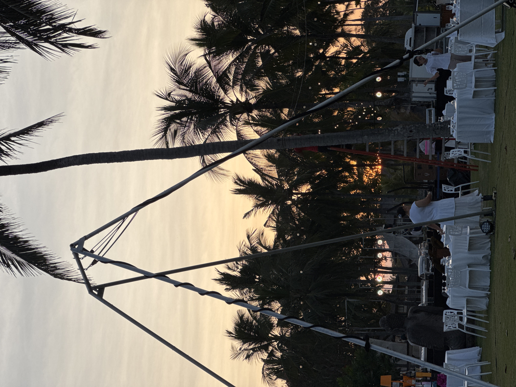 The image depicts an outdoor event setup in Kuta Selatan, Indonesia, at dusk. The scene features a lawn with white tablecloth-covered dining tables and chairs. In the background, a dense line of palm trees silhouetted against an orange and yellow sky suggests the setting is a tropical location. A large statue of Buddha is visible on the left side of the frame, positioned behind some of the tables. Several people are present, with one individual on a ladder near a palm tree, possibly engaged in setting up lights or decorations. Another person stands near a table, interacting with their phone. String lights are strung between trees, contributing to the festive atmosphere. A metal structure, possibly part of a tent or stage rigging, is prominent in the foreground, with cables and supports extending upwards. The overall impression is of a pre-event scene, with preparations underway for a gathering.