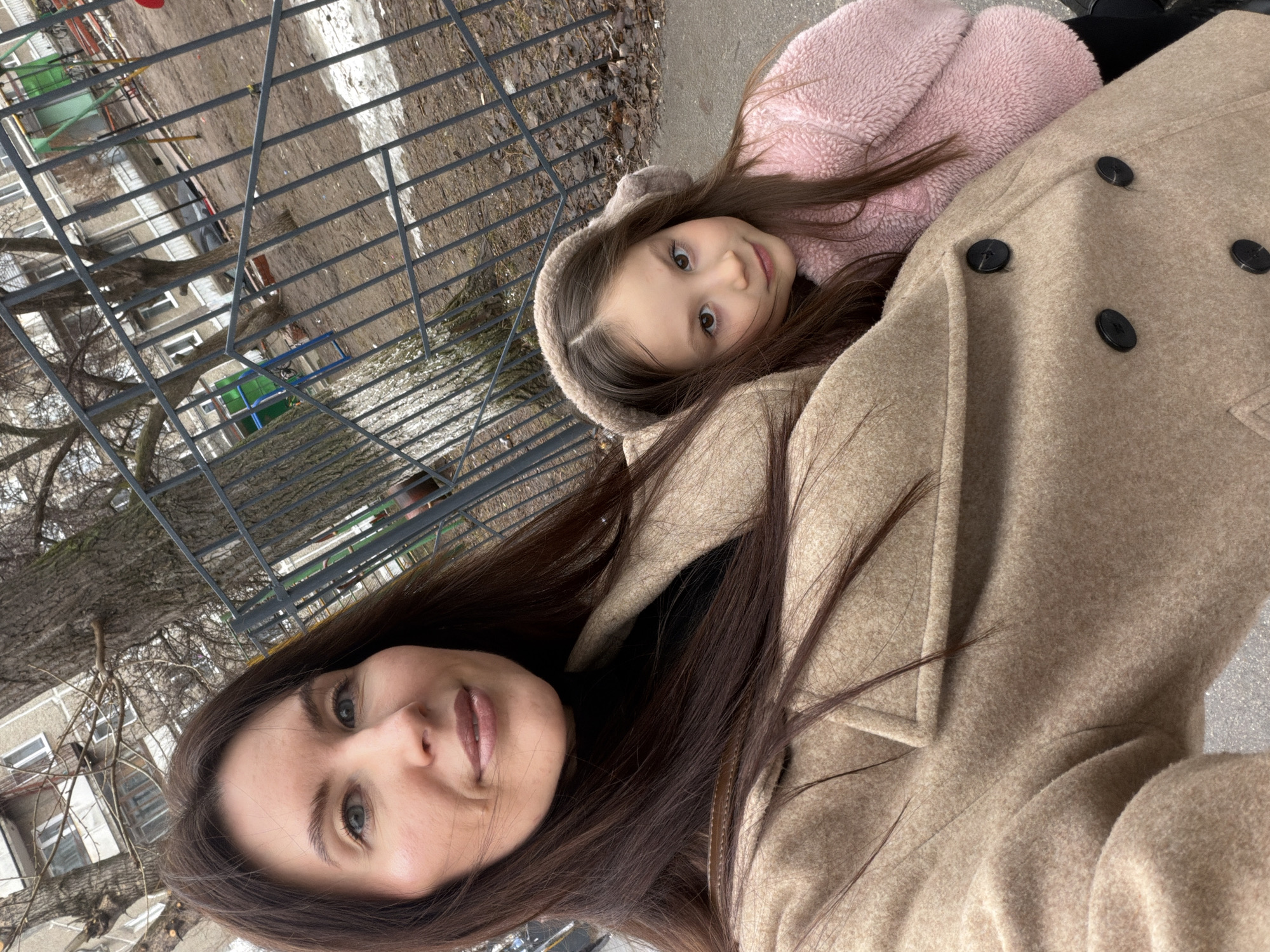 A woman and a child are in a close, selfie-style shot. The woman has long, dark hair and is wearing a tan coat. The child, who appears to be a young girl, is wearing a pink, hooded jacket. They are outside, with a metal fence and a tree visible in the background, and a building. Based on visual landmarks, the location is in Perm, Russia. Patches of snow are visible on the ground.