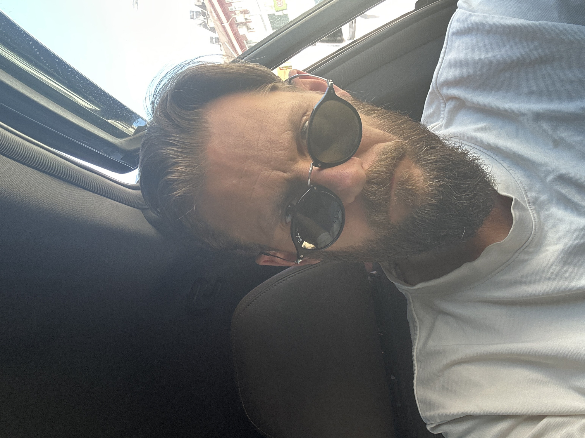 A man with sunglasses is in a car, possibly in Fethiye, Turkey, as suggested by the context. He has dark hair with light brown highlights and a beard. He is wearing a white t-shirt. A car's sunroof is visible at the top left. The car's interior is dark. The man is positioned at an angle in the shot, with his head tilted. The light is bright, suggesting daylight.