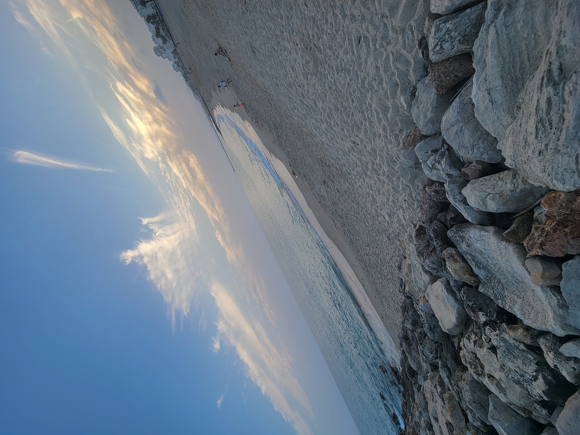 The image shows a wide-angle view of a beach in Quarteira, Portugal, during the golden hour, likely late afternoon or early evening. The sky is a gradient of blues and yellows, with wispy clouds streaking across it. The ocean, a calm turquoise, meets the sandy shore with gentle waves. In the foreground, a rocky embankment lines the edge of the beach. Several people are visible in the distance on the sandy expanse, including a child in orange shorts, and two adults. One adult is playing with a child, and another is lying on a towel. The background features a row of buildings and palm trees, typical of a coastal resort town. The sand is imprinted with small ripples, suggesting a light breeze. The overall atmosphere is serene and recreational.