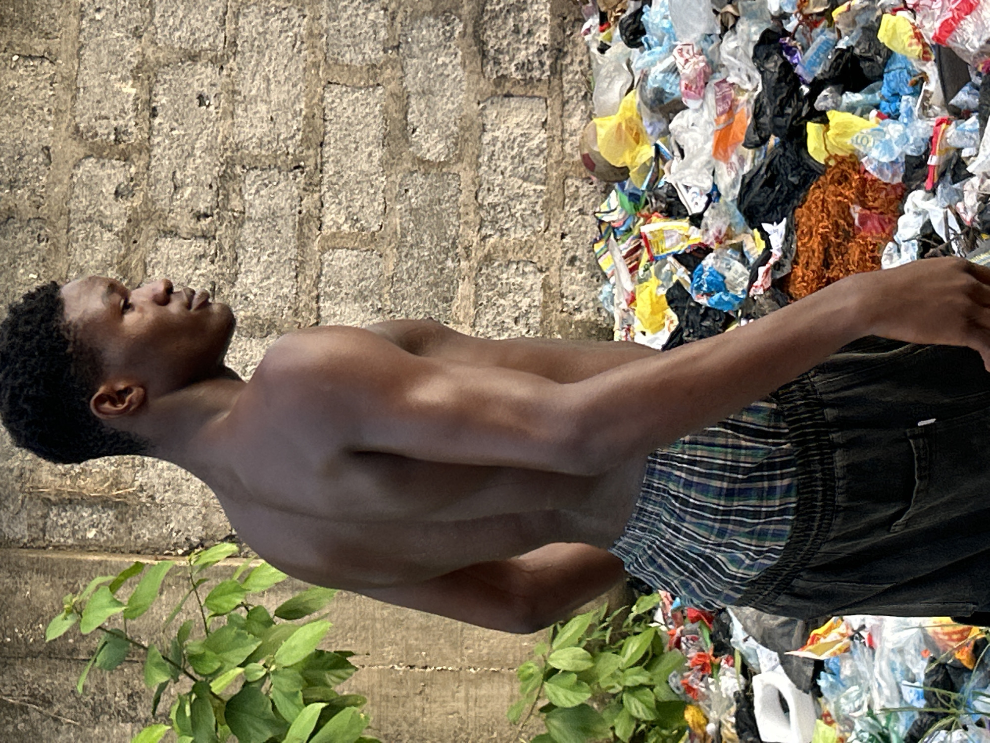 A shirtless young Black man stands among a pile of refuse in Ihiagwa, Nigeria. He is wearing black pants with a blue and green checkered waistband. The refuse includes plastic bags, bottles, and other trash. A concrete block wall is in the background, and green plants are in the foreground.