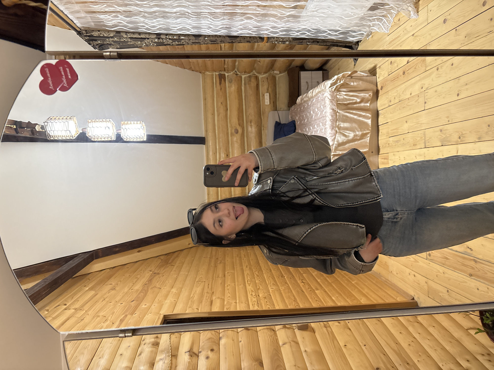 The image shows a young woman in a mirror selfie, likely indoors. She is wearing a gray jacket and jeans, and her sunglasses rest on her head. Her phone is visible in her hand. The background reflected in the mirror suggests a room with wooden walls and floors, characteristic of a log cabin or rustic interior design. A bed with a decorative cover is also visible. The image includes a heart-shaped decoration on the mirror, with a Russian inscription which translates to “Beloved wife." The location is possibly Chaikovsky, Russia, due to prior context.