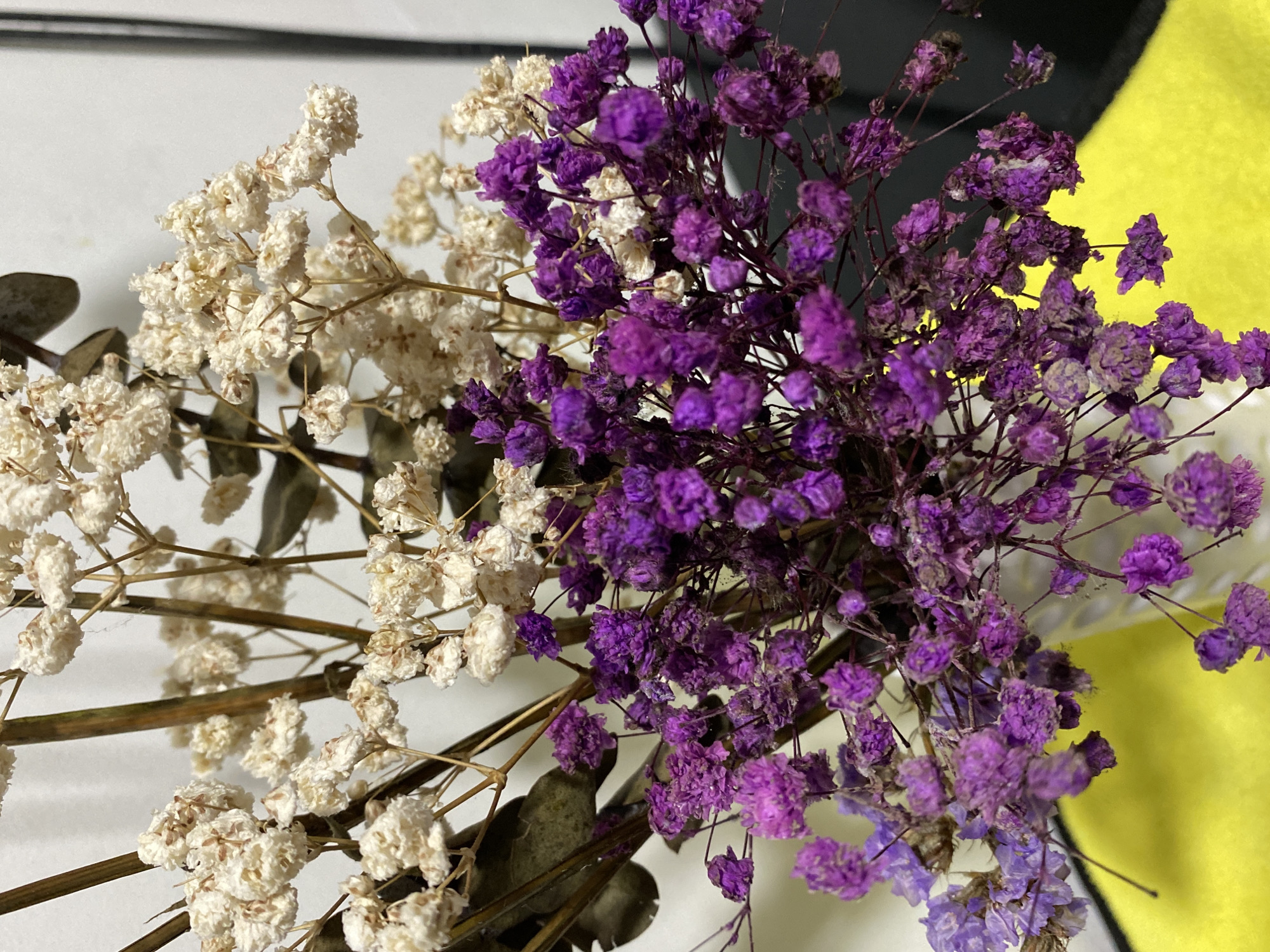 The image displays a close-up view of a dried flower arrangement. The arrangement features clusters of small, delicate white flowers and smaller, vibrantly purple flowers. The stems supporting the flowers are thin and appear to be dried and brown. The background consists of a plain white surface on the left and a dark surface on the right, suggesting an indoor setting. A portion of a bright yellow surface is visible in the lower portion of the image, possibly a tablecloth or a piece of fabric. There are no people or visible actions occurring in the scene. The image provides no information that would suggest a location in Davao City, Philippines, or any other specific geographical location.