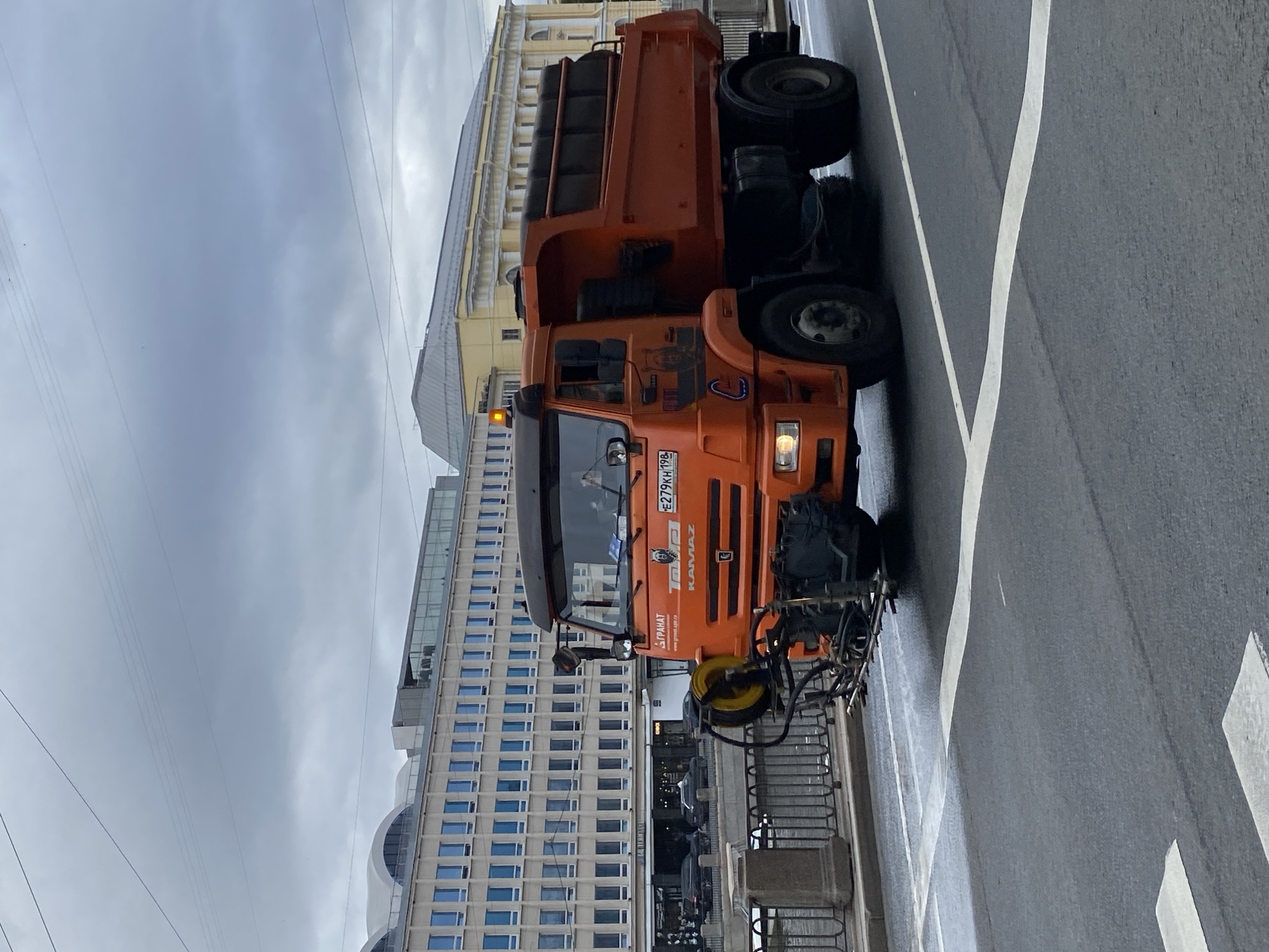 An orange KAMAZ dump truck, equipped with a street sweeping attachment, drives on an overcast day across a street in Saint Petersburg, Russia. The truck is on a bridge, with a canal visible to its left, bordered by a stone embankment and railing. In the background, a large, modern building with a grid of windows stands to the left, while a classical-style yellow building with arched windows is visible to the right. The street is wet, indicating recent rain. The truck's license plate is visible and reads "E279KH 198". There are no visible people in or around the truck.