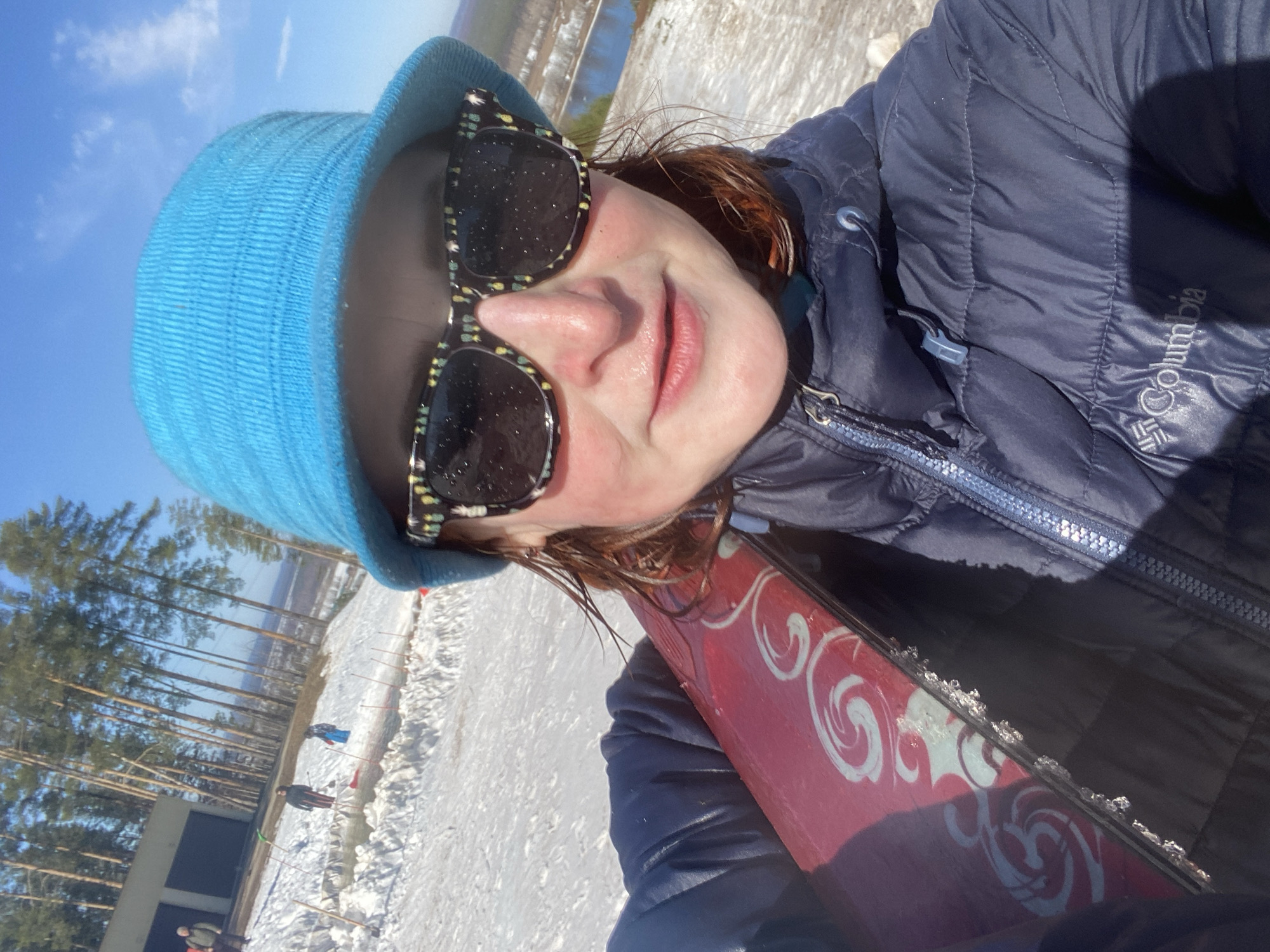 A woman is taking a selfie on a snowy day in Прикамский, Russia. She is wearing a blue hat, patterned sunglasses, and a dark blue Columbia jacket. She is holding a red snowboard with white designs. The background shows a snowy landscape with trees, and a building with people. The sky is clear and blue.