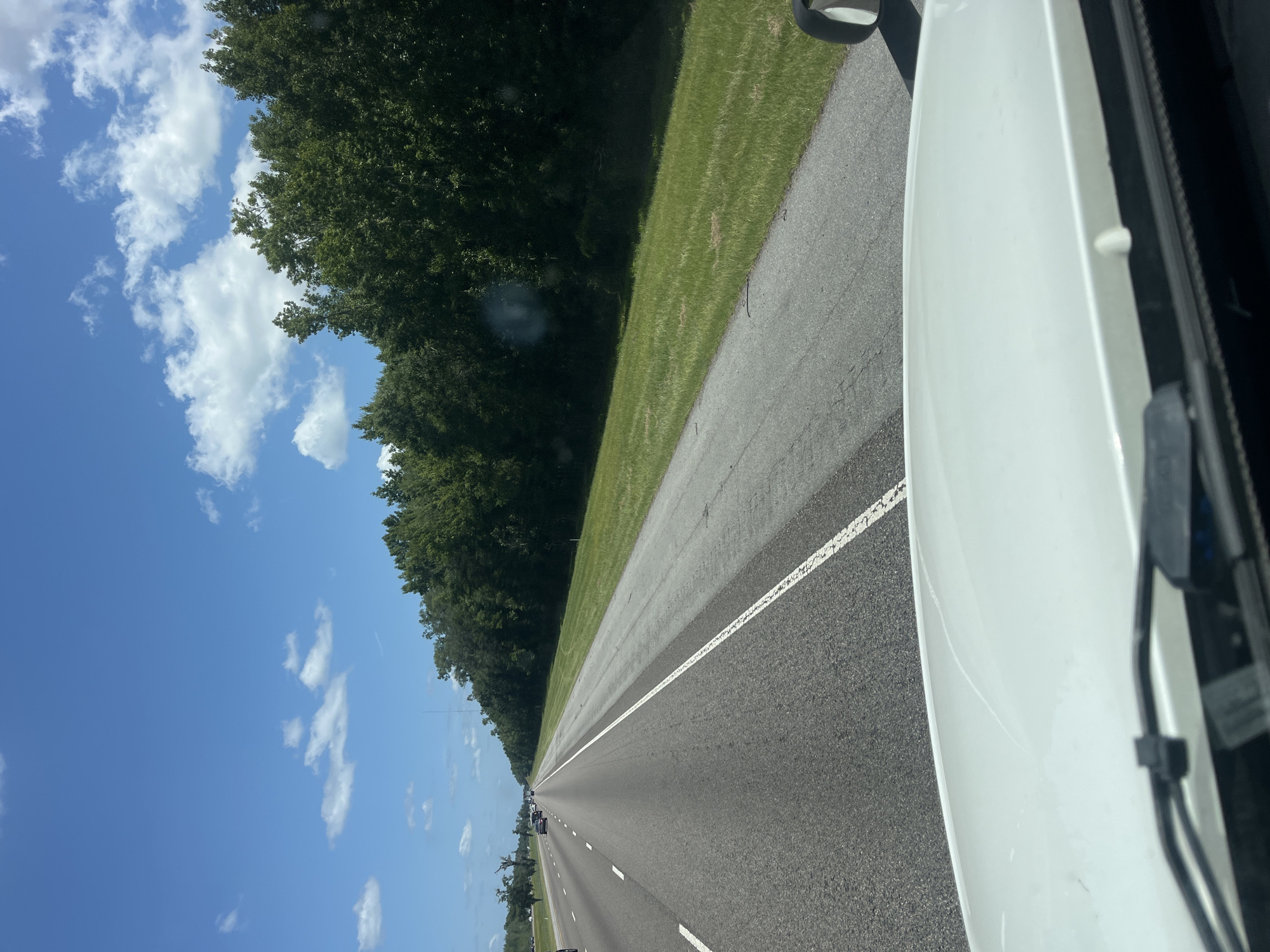 A forward-facing shot from inside a vehicle shows a multi-lane highway in Riceboro, United States, under a bright blue sky dotted with white clouds. The highway stretches into the distance, bordered on the right by a strip of green grass and a dense line of trees. Several cars are visible on the highway. The foreground of the image shows the white hood of the vehicle the photo is taken from, with windshield wipers and part of the dashboard visible.