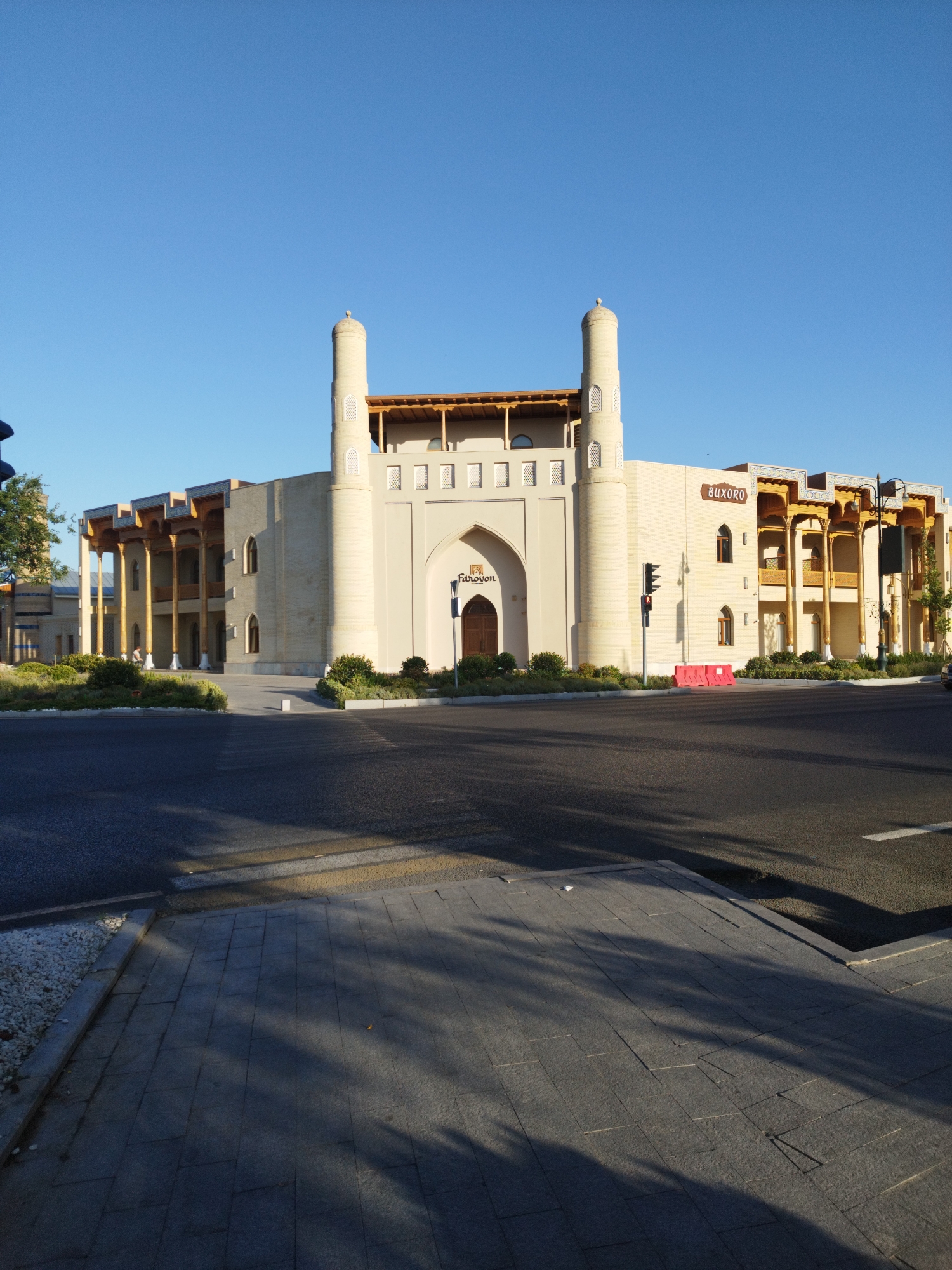 A wide, low-angle shot captures a beige, modern building with two prominent minarets against a clear blue sky. The building features a large arched entrance with the word "Farovon" above it. To the right of the entrance, the building has a sign that reads "BUXORO". The structure is adorned with wooden pillars and decorative eaves, giving it a blend of traditional and contemporary architectural styles. In front of the building, there are neatly trimmed green bushes. A paved walkway with a pedestrian crossing is visible in the foreground, separating the building from the asphalt road. A traffic light is also present. Shadows of tree branches are cast across the walkway.