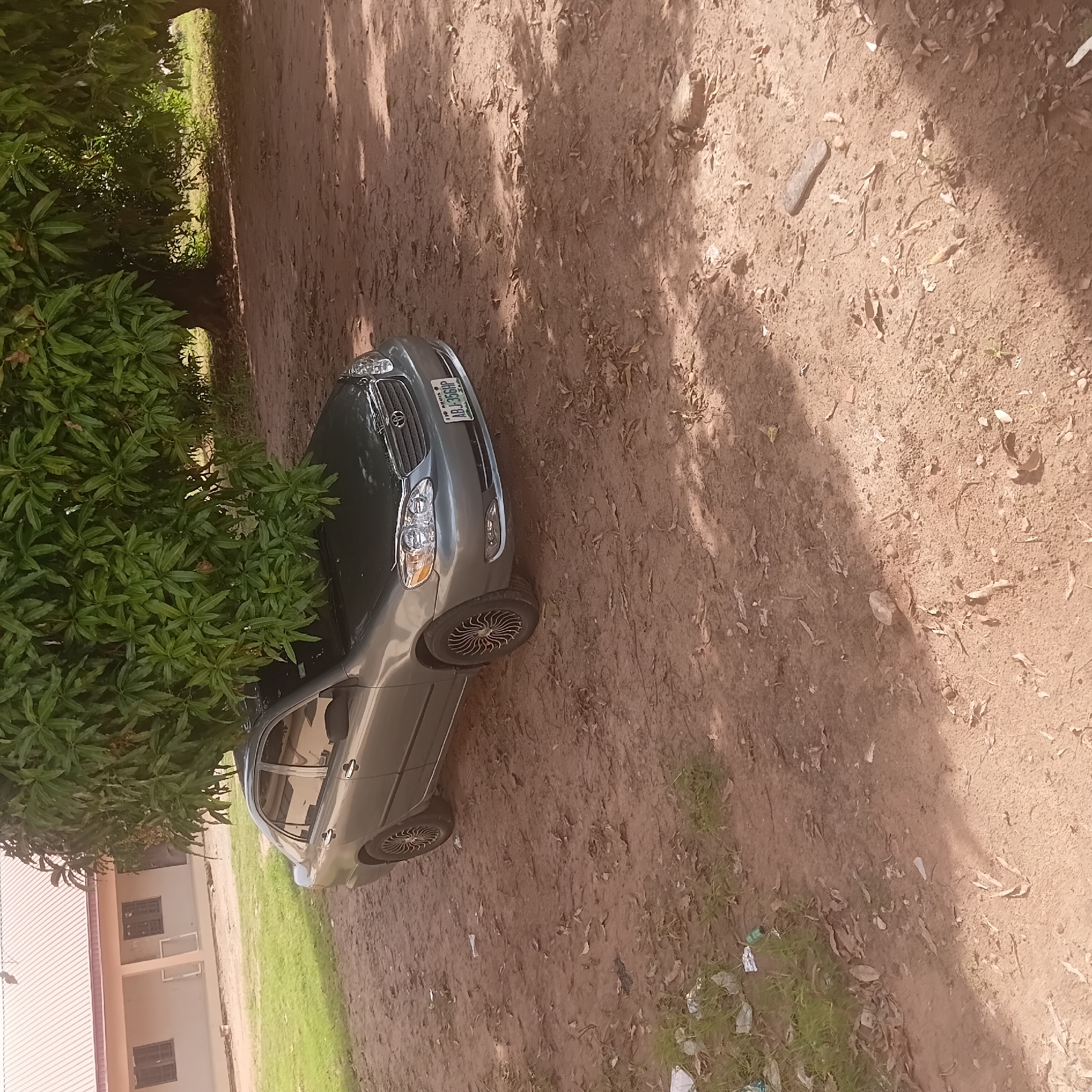 A gray sedan is parked on a dirt ground in front of a building in Wukari, Nigeria. The car is partially obscured by the branches of a large tree. The building appears to be a school or community center, with a light-colored facade and a red-tiled roof. There are a few scattered pieces of trash on the ground. The scene is well-lit by the sun, with shadows cast by the tree.