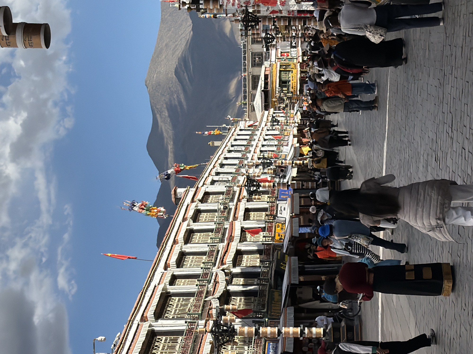 The image captures a bustling street scene in Chengguan District, China, characterized by traditional Tibetan architecture and a vibrant flow of people. The perspective looks down a paved street lined with multi-story buildings, backed by a large, barren mountain under a clear blue sky with scattered clouds.
The main subjects are numerous people walking along the street, many of whom appear to be engaged in a procession or pilgrimage. They are dressed in a mix of traditional and modern attire. Along the side of the street, attached to the buildings, are rows of cylindrical prayer wheels. Several individuals are seen actively turning these prayer wheels as they move, indicating a religious or cultural practice.
The buildings exhibit distinctive Tibetan architectural elements, with white facades, dark-framed windows, and decorative cornices. Colorful prayer flags, typical of Tibetan culture, are strung up in various locations, adding to the cultural atmosphere. A red flag, possibly featuring a single star, is also visible flying from a pole on one of the buildings.
Visible text includes characters on some of the prayer wheels. On one set, Chinese characters like "右" (yòu - right) and "上" (shàng - up) are discernible. On other prayer wheels, Tibetan script, including what appears to be "ཀ་" and "ཁ་", can be seen. Signs with illegible text are also present on the building facades.
The scene takes place on a bright, sunny day, likely in the late morning or early afternoon, given the clear illumination and the quality of the shadows. The majestic mountain range in the background firmly places the setting in a high-altitude, mountainous region, consistent with the Tibetan Plateau.