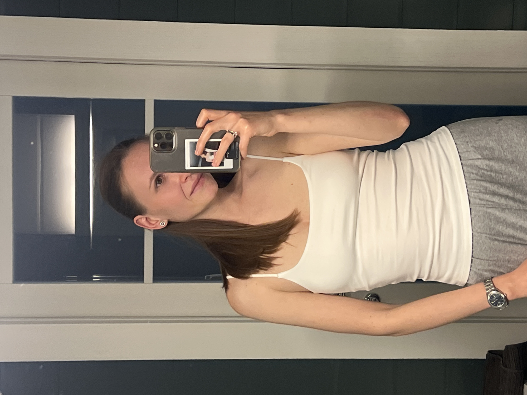 A woman is taking a selfie in a bathroom. She is wearing a white tank top and gray pants, and she has brown hair in a ponytail. She is holding a phone in her hand, and the phone has a sticker on the back. She is wearing a watch on her left wrist. The bathroom has a white door and a window with a blue wall visible through it. There is no evidence in the image of the location being in Khimki, Russia.