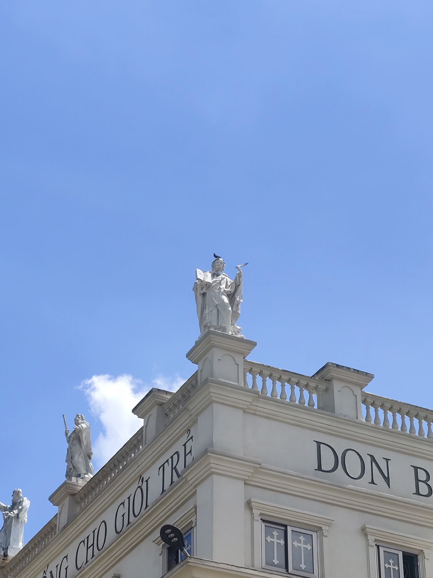 The image captures the upper facade of a white building against a clear blue sky with a few wisps of clouds. Several white statues of robed figures are positioned along the rooftop. One prominent statue in the center holds an open book, and a small black bird is perched on its head. Another statue to the left holds a staff. Text is visible on the building's facade: "NG CHO GIỚI TRẺ" is written in large, dark letters across the left side, and "DON B" is visible on the right. Two windows with white cross patterns are also present on the lower part of the facade. This building is identified as Don Bosco Da Lat, located in Da Lat, Vietnam.