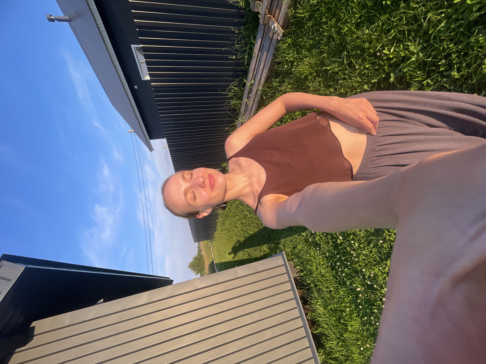 A young woman with blonde hair pulled back, wearing a brown crop top and gray pants, stands in a grassy yard in the village of Podshivalovo, Russia. She is taking a selfie with her right arm extended, holding the camera. Her eyes are closed, and she has a slight smile. The sun is low in the sky, casting a warm glow and long shadows. To her left is a gray, modern-looking building with a sloped roof. To her right is a black metal fence and a dark-colored building with a sloped roof, a chimney, and a white-framed window. In the background, there are trees and a bright blue sky with thin white clouds. A pile of wooden planks lies on the grass near the fence.