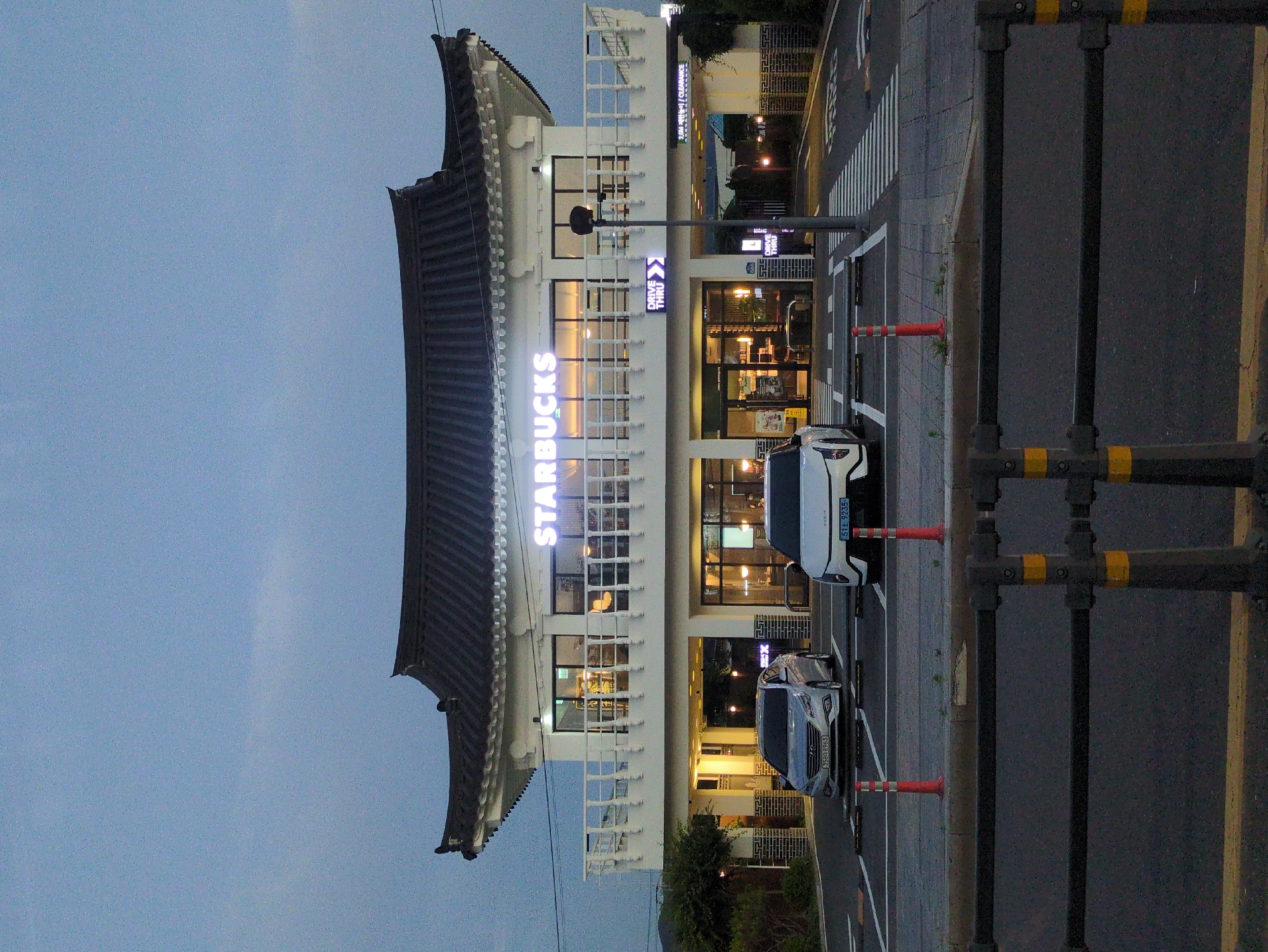 The image depicts a Starbucks store in Gyeongju-si, South Korea. The building is a two-story structure with a traditional Korean-style roof. The Starbucks logo is illuminated on the exterior. Cars are parked in a designated parking area. A street is visible next to the store, with a pedestrian crossing. The sky is visible in the background, suggesting it is dusk or dawn. A dark-colored metal guardrail is in the foreground, with orange reflective striping.