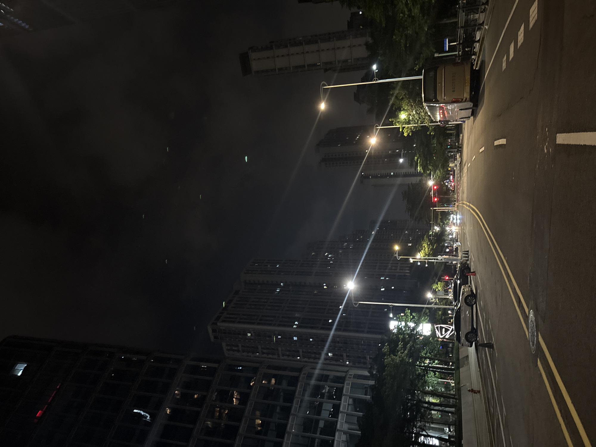 The media file depicts a night scene on a street in Haizhu District, China. The perspective is from the side of the road, looking down the street towards a cluster of tall buildings. The time of day is clearly night, indicated by the darkness and the streetlights illuminating the scene. The weather appears to be slightly hazy or foggy, as the lights from the buildings and streetlights are diffused.
The main subjects include several high-rise buildings with illuminated windows, suggesting they are residential or office complexes. Trees line one side of the street, their foliage lit by the streetlights. A road with distinct yellow and white lane markings is visible, with parked vehicles along the side, including a bus and several electric scooters. The road is wet in places, possibly from recent rain.
There is no overt activity or event taking place, but the presence of parked vehicles and illuminated buildings suggests a normally active urban area at rest during the night. Notable details include the style of the buildings, which appear modern. The streetlights are contemporary, with some featuring a curved design.
Visible text is limited to a license plate on the bus, which appears to be Chinese characters and numbers. The overall emotion conveyed is one of urban tranquility and the quiet hum of a city at night. The scene is well-lit but also hints at the atmospheric conditions due to the diffusion of light.