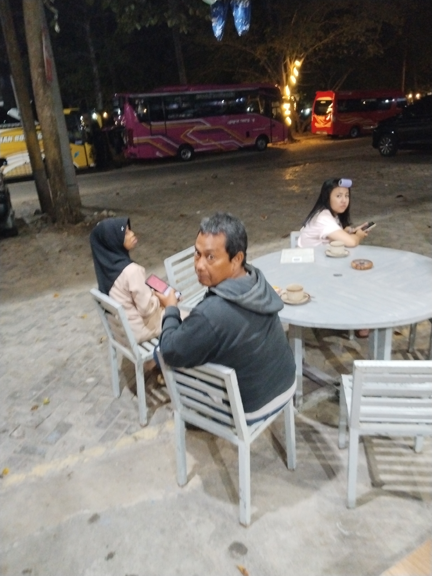 In the city of Prigi, Indonesia, a group of people are gathered outdoors at night. In the foreground, a man in a gray hoodie is seated, facing the camera. To his left, a woman wearing a black hijab and a light-colored top is seated, holding a pink phone. A round table is positioned in front of them, with two cups and saucers on it. Further in the background, a young woman with hair rollers is seated, also looking towards the camera and holding a black phone. Behind the people, several buses are parked. One bus is pink and decorated with graphics, while another is red. A black car is also visible parked to the right of the red bus. The environment appears to be a paved area with some trees and lights in the background.