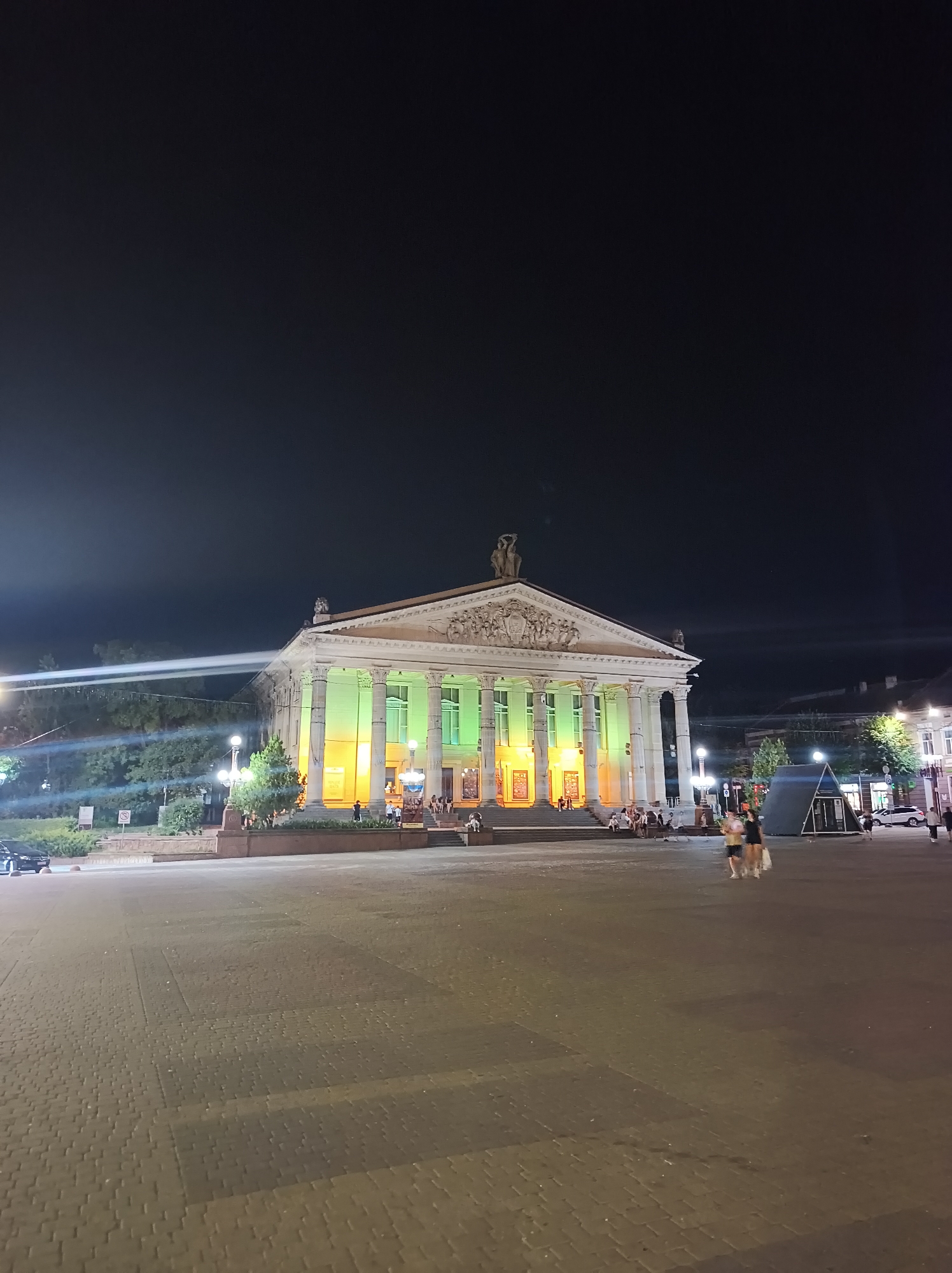 The image depicts Ternopil Theatre, also known as Ternopil Academic Regional Ukrainian Drama Theatre, at night. The neoclassical building features a prominent facade with large columns and ornate sculptures. The theater is illuminated with green and yellow lights, creating a vibrant contrast against the dark night sky. In the foreground, a paved plaza is visible, with a few people walking and standing around. Some trees and streetlights are also present. The overall scene suggests a lively evening atmosphere in the city center.