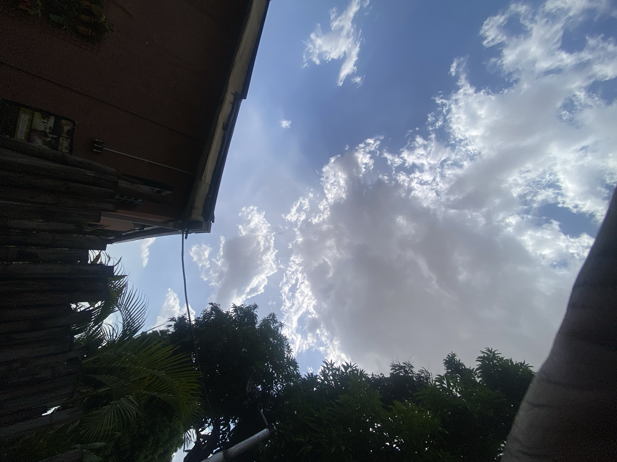 The image, taken from a low angle, shows a view of the sky with scattered clouds. In the foreground, there are tree branches with green foliage, and a section of a building. The building is partially visible on the left side, with a section of brown wall and an overhanging roof. The sky is blue with varying densities of white and gray clouds. The time of day appears to be during daylight. The location is not discernible from the image, and no elements of the city of Barden Yana, Nigeria are present in the image. There are no people visible in the photograph.