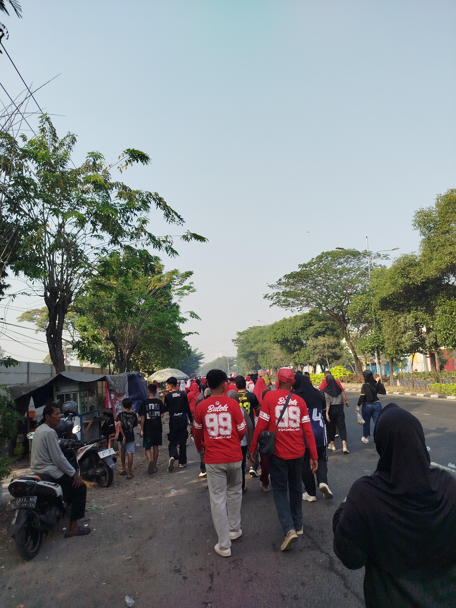 A group of people in red shirts are walking down a street in Surabaya, Indonesia. The sky is clear and the sun is shining. There are trees on either side of the street, and a fence in the background. A man is sitting on a motorcycle on the left side of the street, and a few other people are walking in the distance. The people in red shirts appear to be part of a group or event, as they are all wearing similar attire. Some of the red shirts have the number "99" on them, and some have text that is not fully legible. The overall atmosphere seems to be one of a casual gathering or procession.