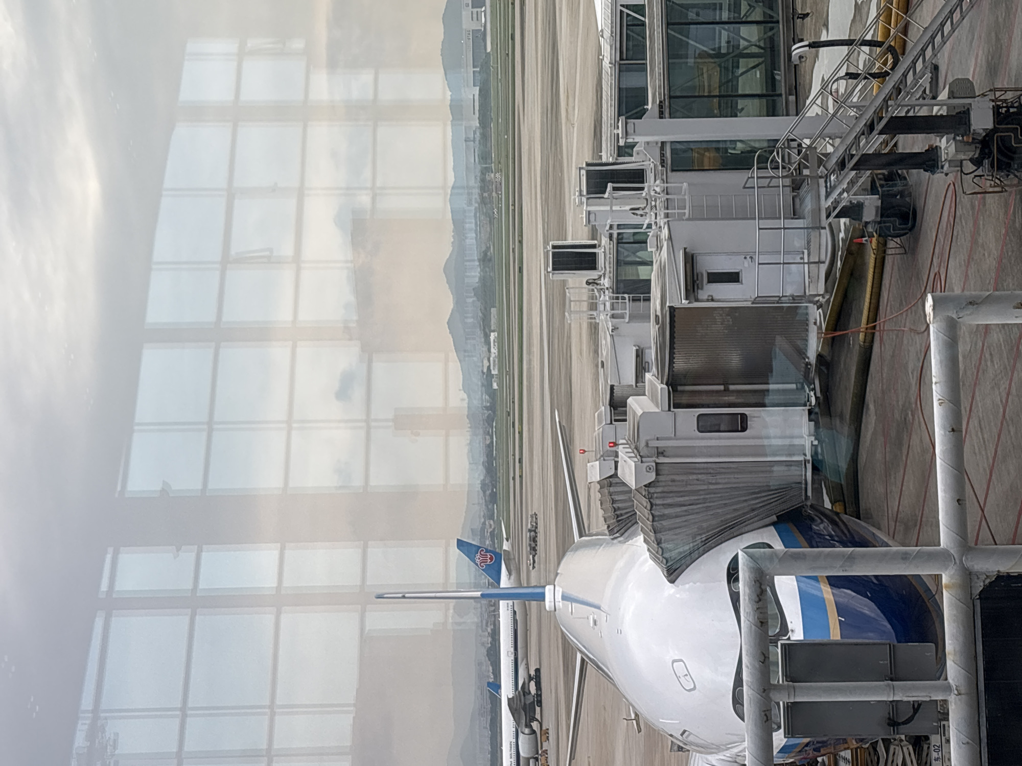 The image appears to be taken from inside an airport terminal, looking out through a window. A passenger boarding bridge is connected to an airplane with blue and gold stripes, possibly ready for boarding or deplaning. In the background, mountains are visible in the distance. The window has a grid-like pattern due to its construction. The location of the image is unlikely to be in Khimki, Russia, because the airline colors do not belong to a Russian airline, and the background is more likely a coastal or mountainous landscape, not a flat terrain.