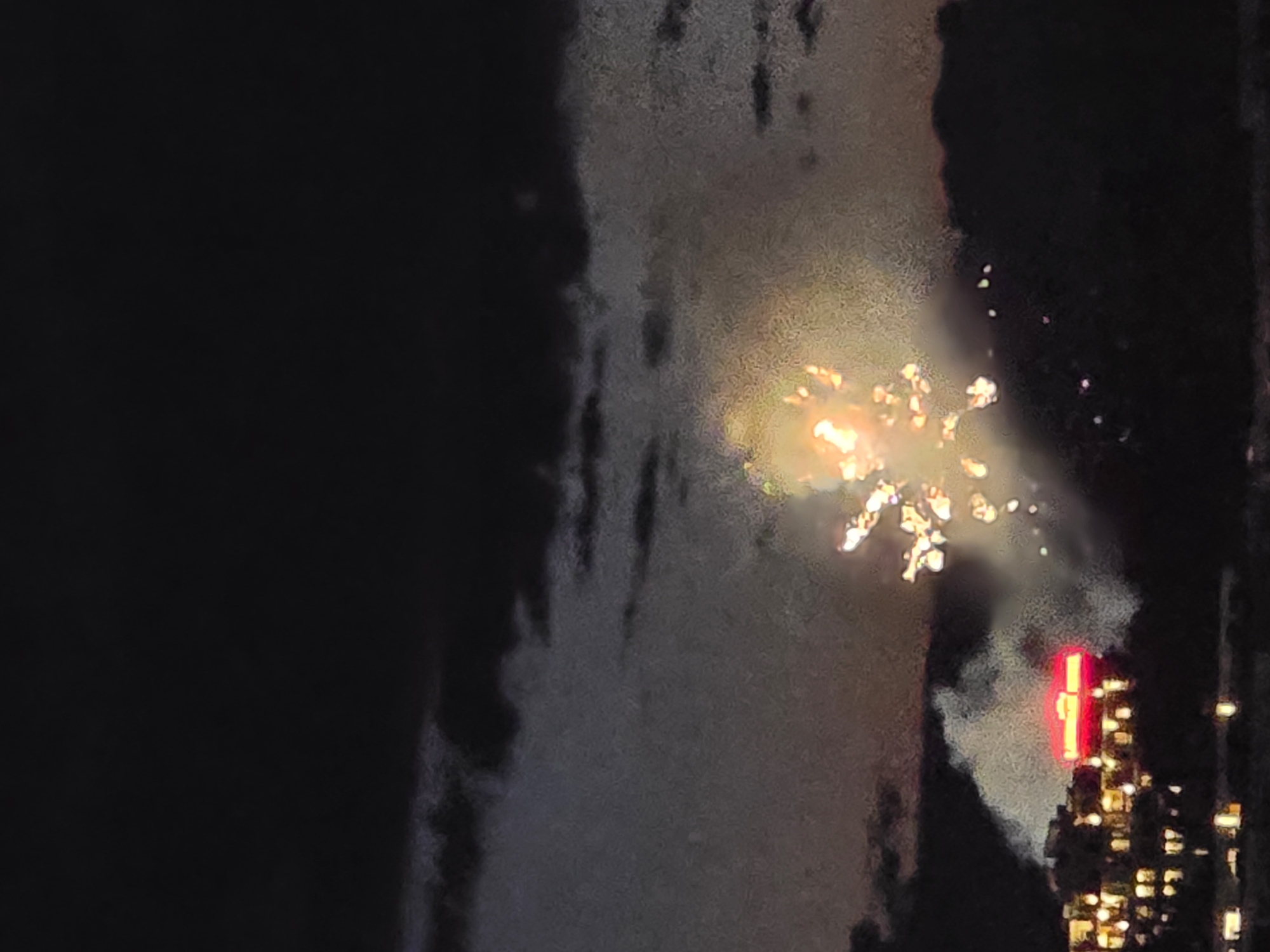Captured at night, the photograph presents a view of fireworks exploding in the sky over what appears to be a city skyline. The fireworks exhibit a bright, multi-faceted pattern with light trails emanating outwards. Below the fireworks, building lights are visible, including what appears to be a brightly lit sign or structure emitting a red glow. The surrounding environment is dark, creating a stark contrast between the bright bursts of the fireworks and the night sky. The exact location and city are not identifiable based on the provided information.