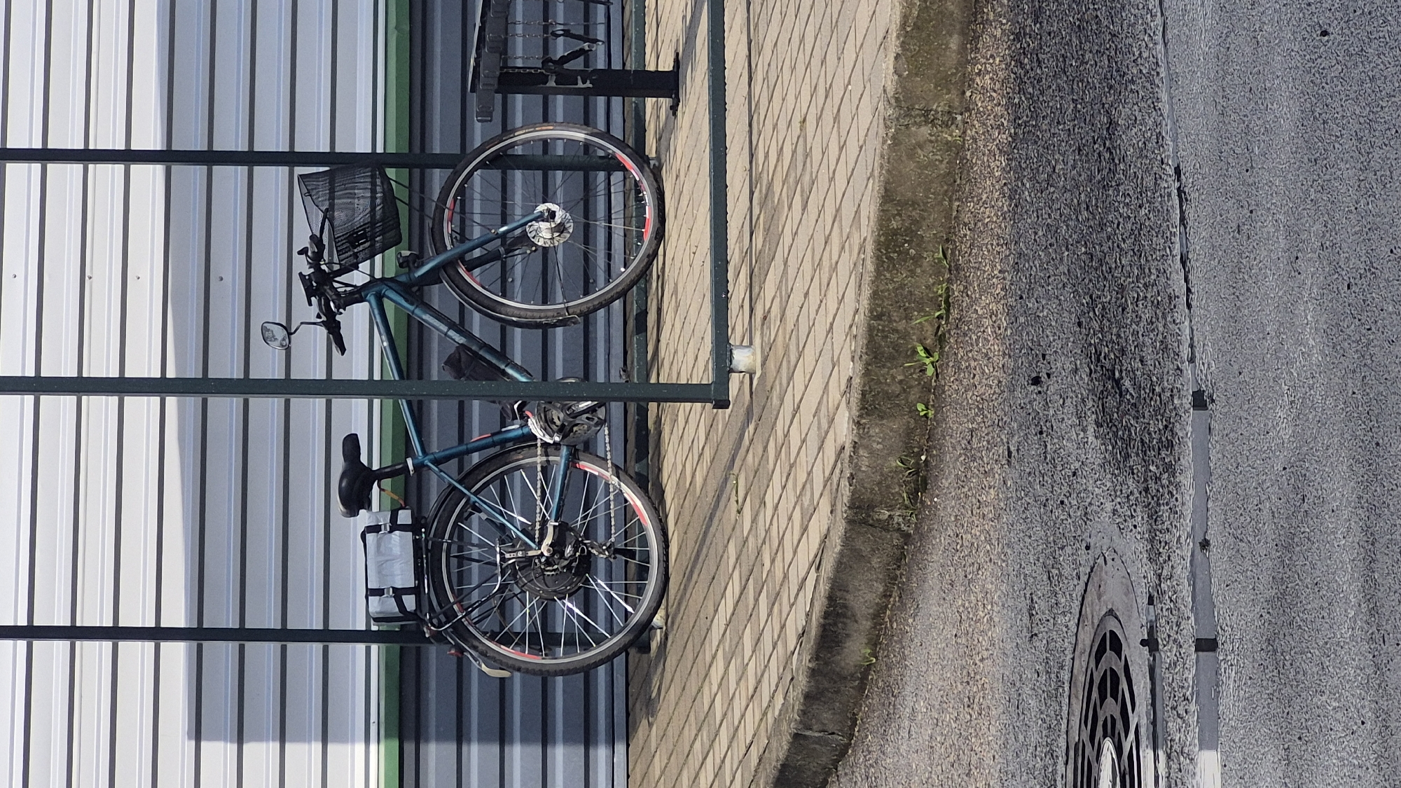 A teal bicycle is parked under a metal shelter with a corrugated metal wall behind it. The bicycle has a basket on the front and a rack on the back with a rectangular white bag. The bicycle is on a paved walkway made of interlocking bricks. In the foreground, there is a curb and a street with a manhole cover. The scene appears to be in an outdoor urban setting, possibly in Pabradė, Lithuania. There are no people visible in the image.
