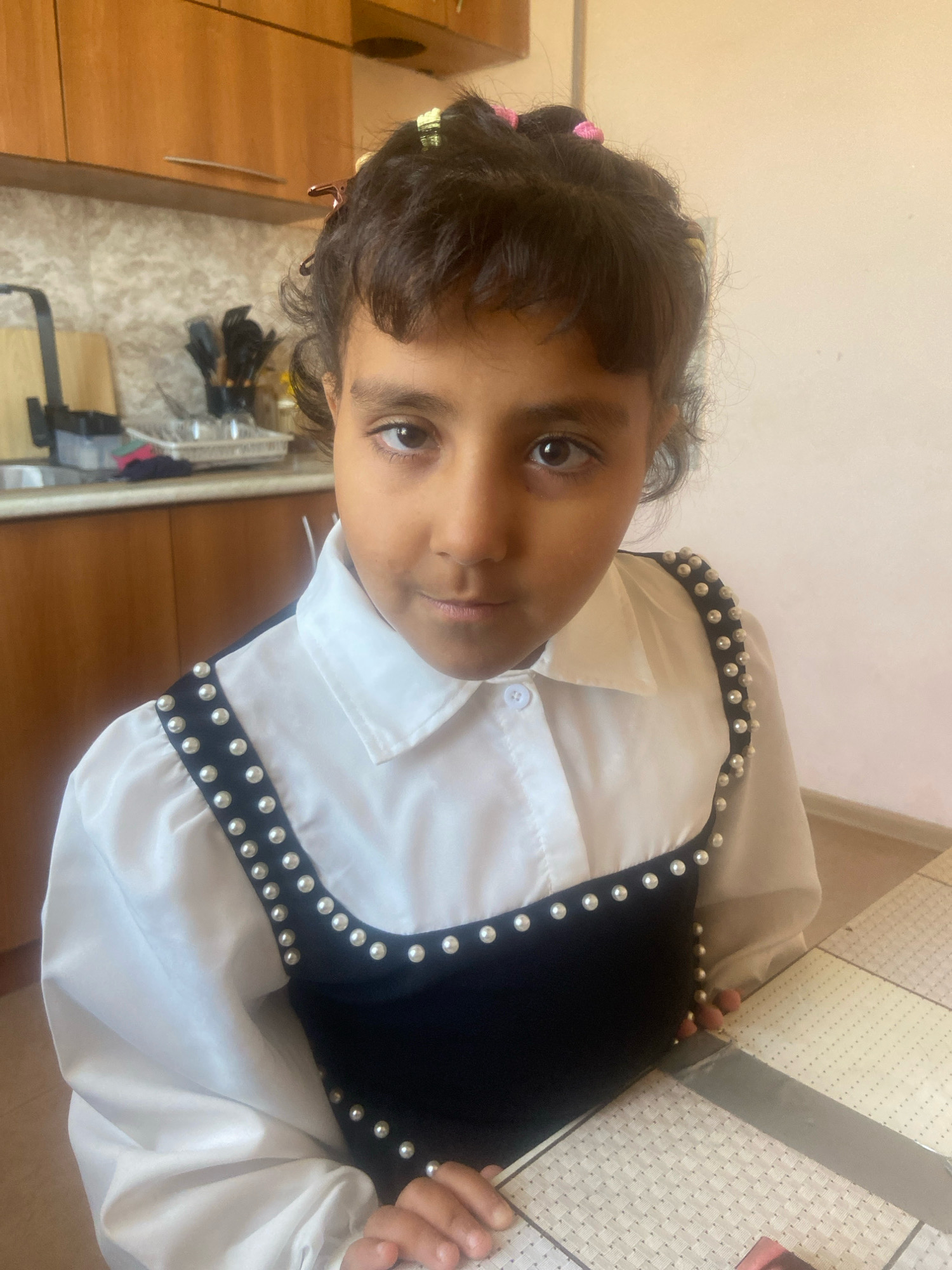 A young girl with dark hair and brown eyes is seated at a table in what appears to be a kitchen. She is wearing a white collared shirt with a black sleeveless overlay adorned with pearls. Her hair is styled with pink and copper hair clips. In the background, wooden cabinets are visible above a countertop with kitchen utensils. The table surface has a patterned texture. The lighting suggests an indoor environment.