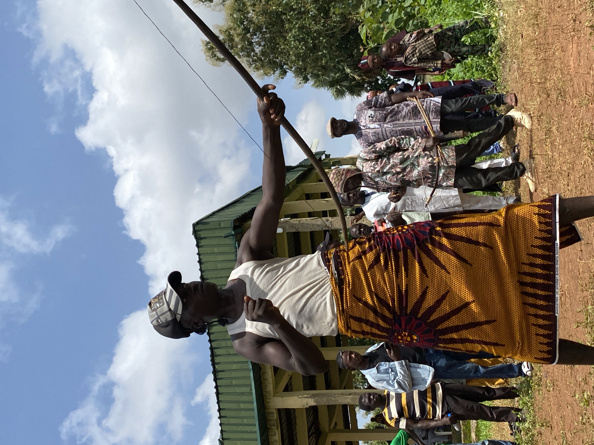 The scene depicts a man in Maigizo, Nigeria, dressed in a white tank top, patterned hat, and yellow and maroon patterned cloth wrapped around his waist. He is holding a long, thin, dark brown rod or pole overhead, possibly demonstrating or performing a ritual. Behind him, a group of men stand, some wearing traditional clothing and hats. The background features a low, green-roofed structure and lush green foliage. A blue sky with scattered clouds is visible above. The ground appears to be a mix of dirt and grass. The presence of people dressed in traditional attire suggests a cultural event or gathering.