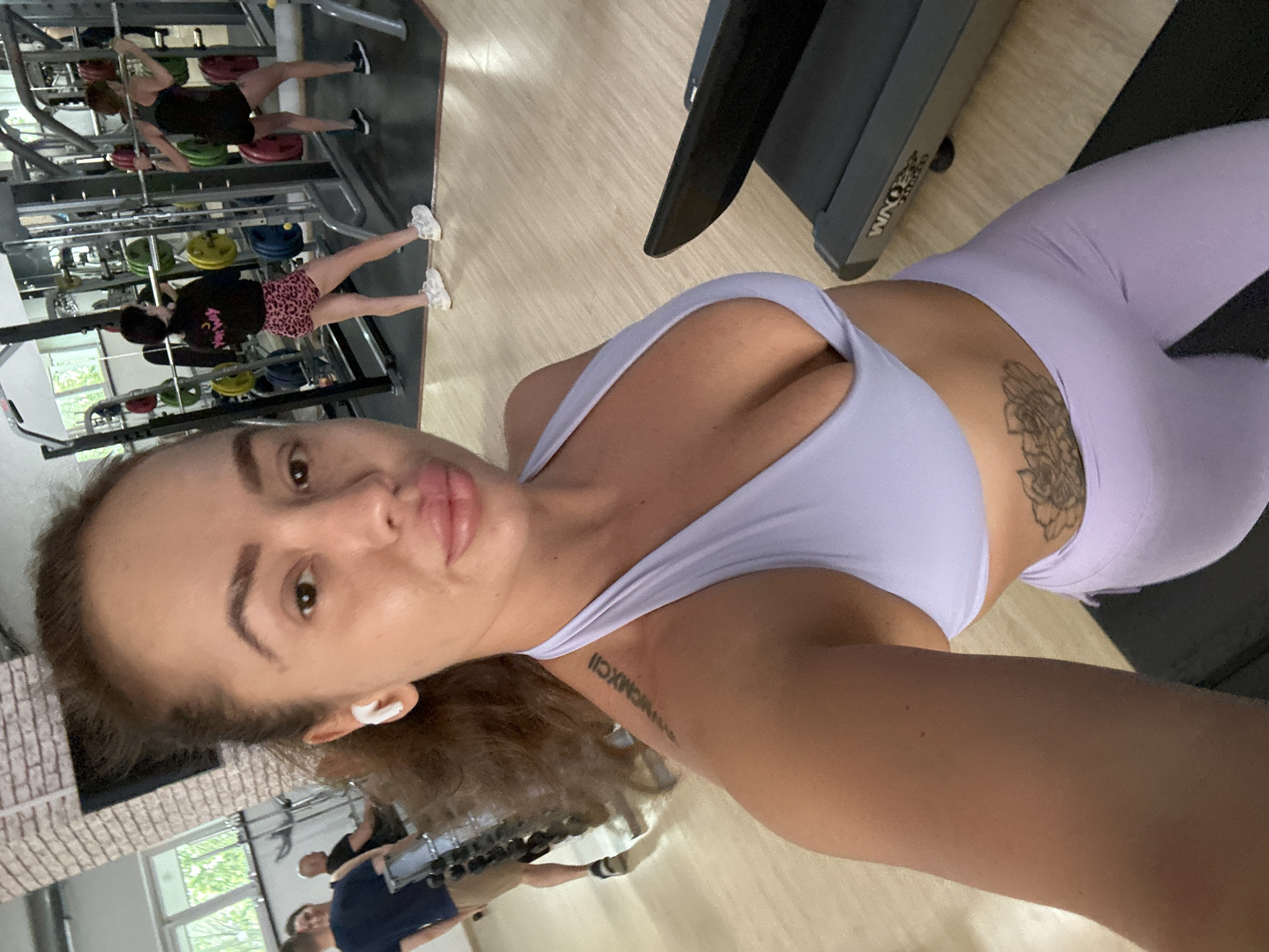 In a gym setting, a woman with brown hair and light skin takes a selfie. She is wearing a light purple sports bra and matching leggings. She has on white wireless earbuds. A tattoo of Roman numerals is visible on her left shoulder, and a floral tattoo is visible on her right hip. To her right, a treadmill is partially visible. In the background, two other women are using weightlifting equipment, one of whom is wearing pink leopard print shorts. A man in a navy blue shirt and tan shorts is standing to the left, facing away from the camera. The gym has brick walls and a wooden floor. The image does not contain any specific information to indicate it is located in Yuzhnouralsk, Russia.