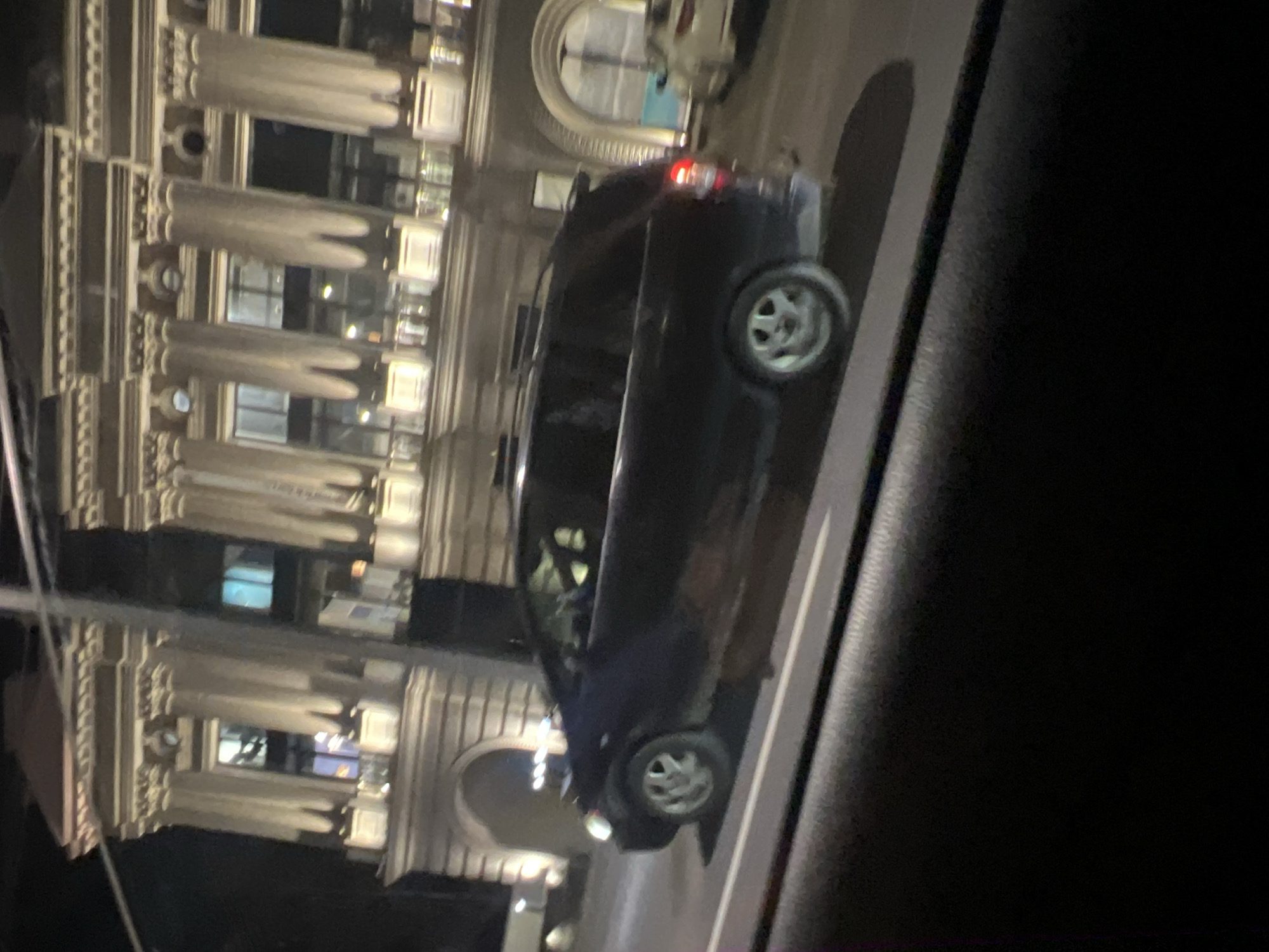 A night shot taken from inside a vehicle shows a black car on a street in Yerevan, Armenia. The car is driving past a large building with prominent columns and arched windows. The building is illuminated, showcasing its architectural details. The interior of the car the picture is taken from is dark, with the side window visible. The street is also dimly lit, but the building provides the primary source of light in the scene.