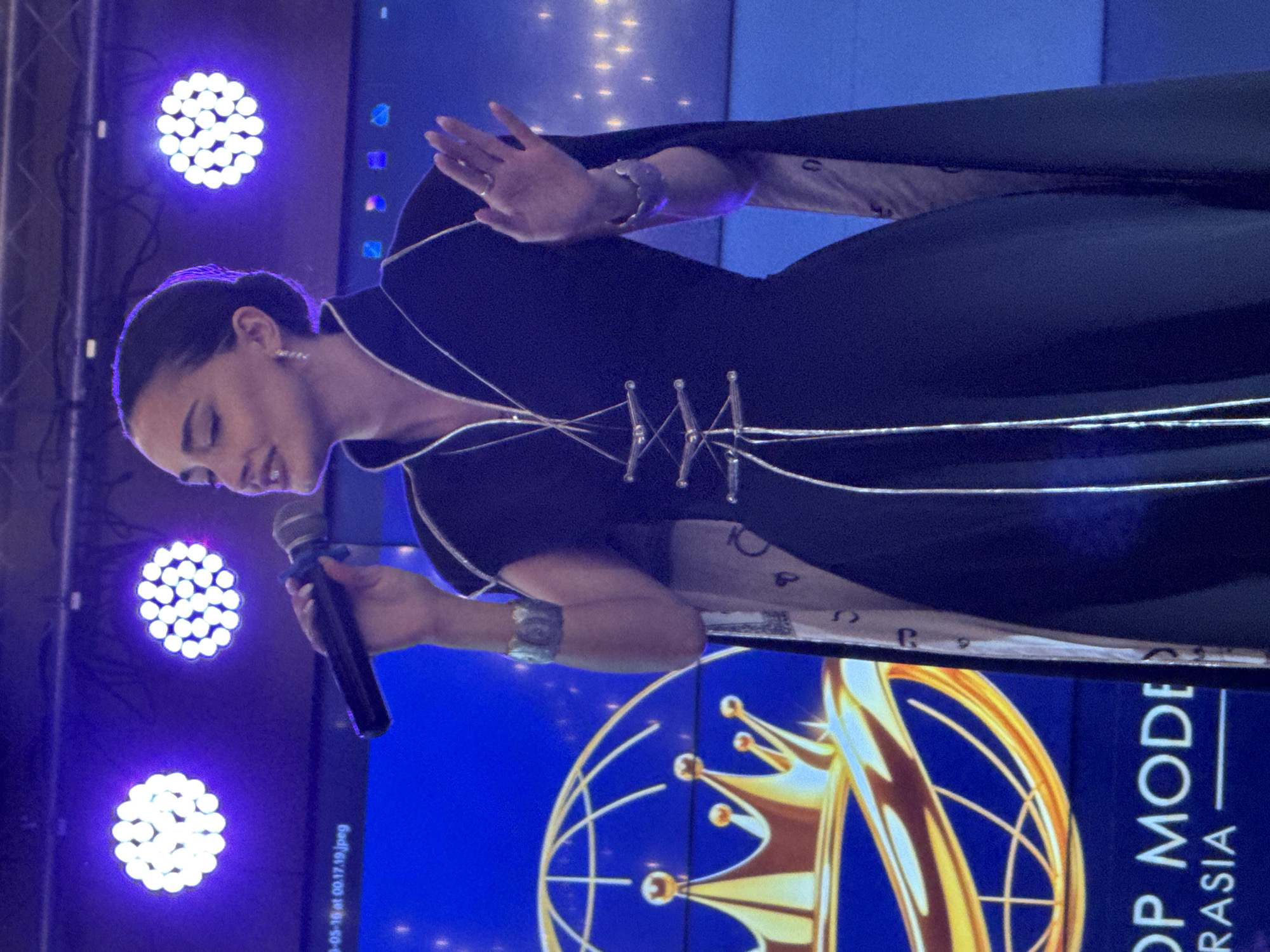 A woman is speaking into a microphone in front of a screen that reads "Top Model Eurasia". She is wearing a dark dress with silver trim. The background features stage lighting. The location may be Batumi, Georgia.