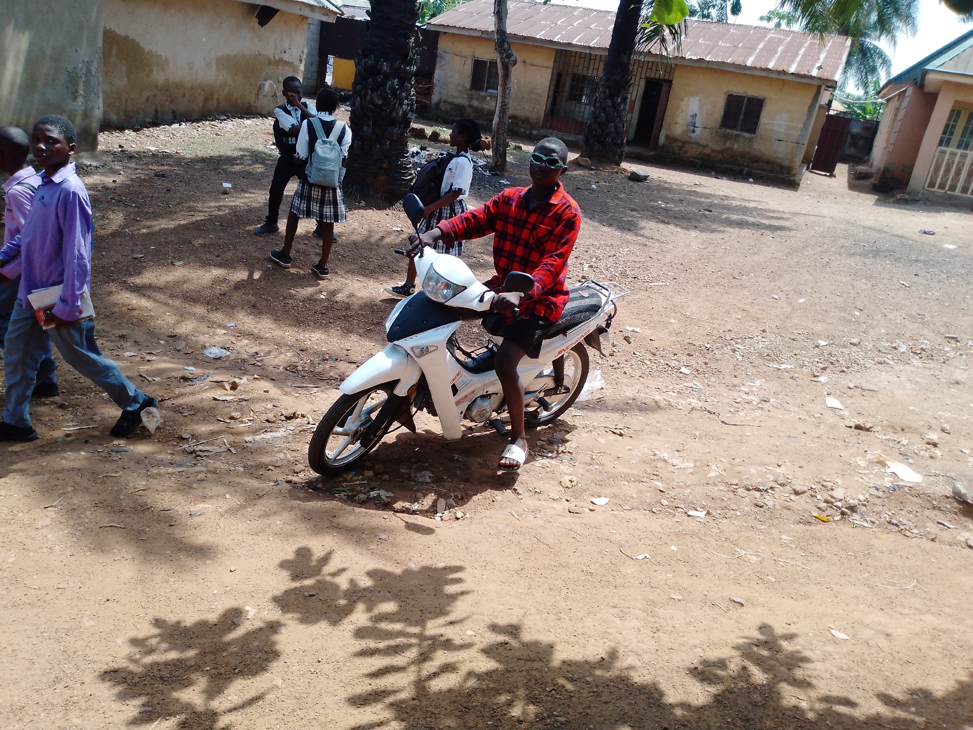 A young man is sitting on a white motorcycle in a dirt lot in an unknown location, city Unknown. He is wearing a red and black plaid shirt, black shorts, sunglasses, and white sandals. There are several people, seemingly school children in school uniforms, walking nearby. The background contains weathered buildings with corrugated metal roofs and tall palm trees.