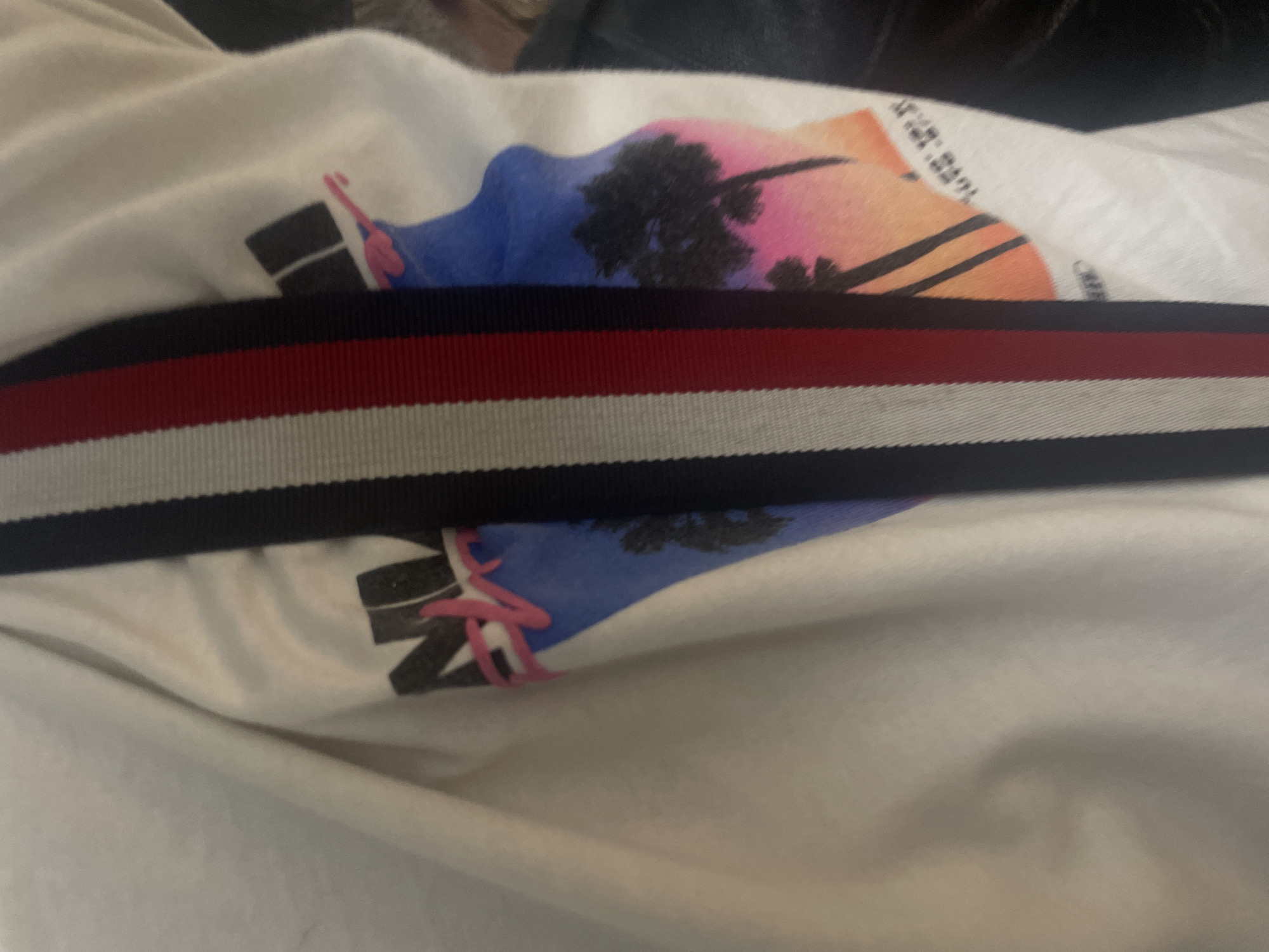 The media file shows a close-up of a white garment, possibly a t-shirt or sweatshirt, laid out on a surface indoors in Yerevan, Armenia. No people are visible.
A prominent feature is a horizontal ribbed trim positioned across the garment. This trim displays distinct stripes in navy blue, red, and white.
Above and below this striped trim, a colorful graphic print is visible on the white fabric. The graphic illustrates a sunset or sunrise scene with a gradient sky transitioning from blue on the left, through pink in the center, to orange and yellow on the right. Silhouettes of palm trees are depicted centrally within this vibrant sky.
Visible text within the graphic is partially obscured and fragmented. On the upper right side of the graphic, above the orange segment, black letters appear to spell "NEW YORK" or a similar phrase. On the left side of the graphic, within the blue area, bold black letters are visible, including what resembles an 'N', accompanied by a thin, curved pink design element.
The garment is slightly creased, indicating its soft fabric texture. The background is out of focus and dark, suggesting a darker surface or fabric beneath. The image captures a still life, focusing on the garment's design and color scheme.