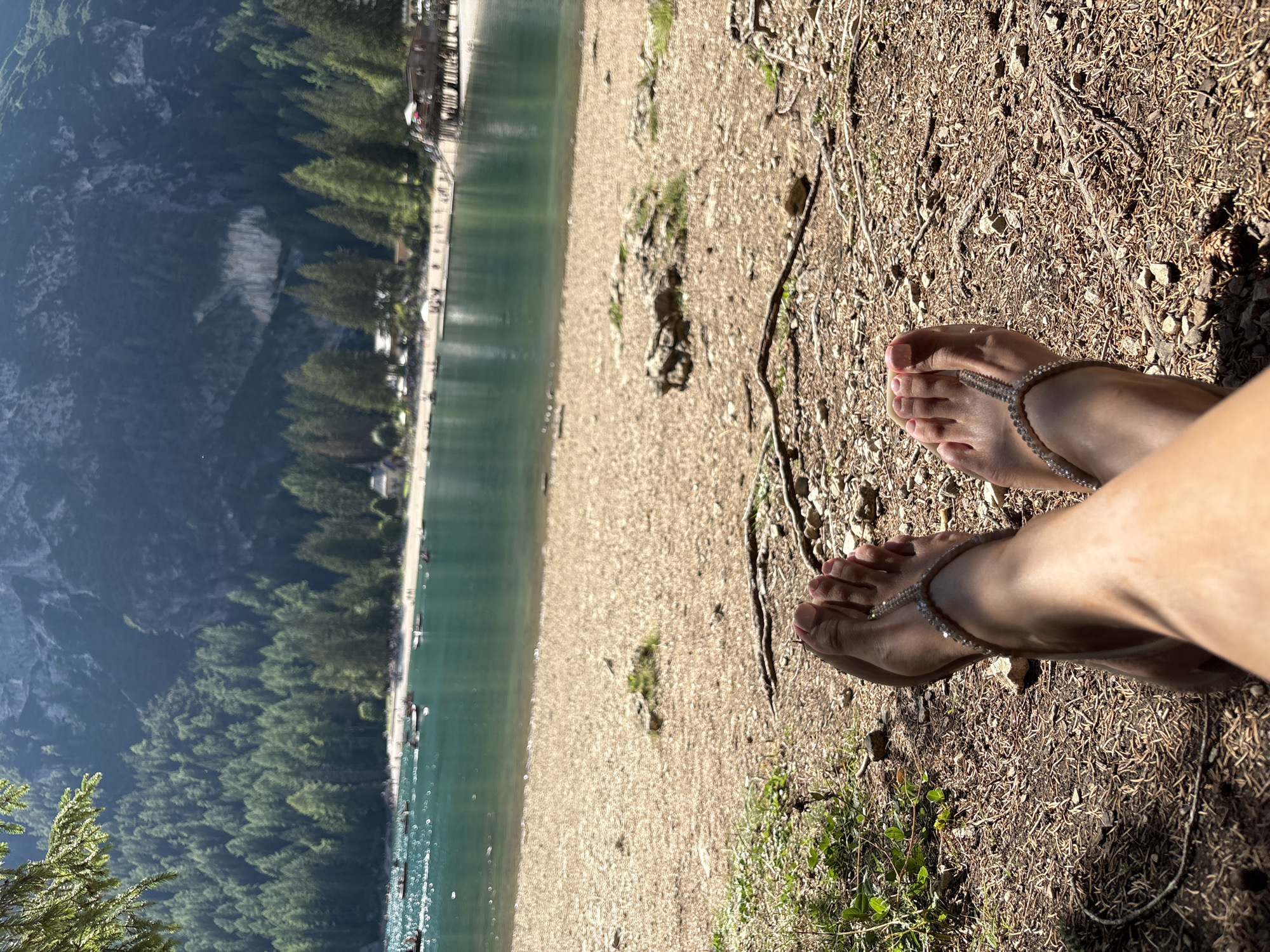 The image shows a first-person perspective of a person's legs and feet resting on the ground near the shore of a lake. The feet are adorned with jeweled sandals. In the background, a body of water with several small boats is visible, along with a sandy beach populated by many people. A dense forest of pine trees covers the slopes of a large mountain range that dominates the horizon. The scene appears to be the lake in Prags - Braies, Italy, a popular tourist destination known for its natural beauty. The lighting suggests it is a sunny day.