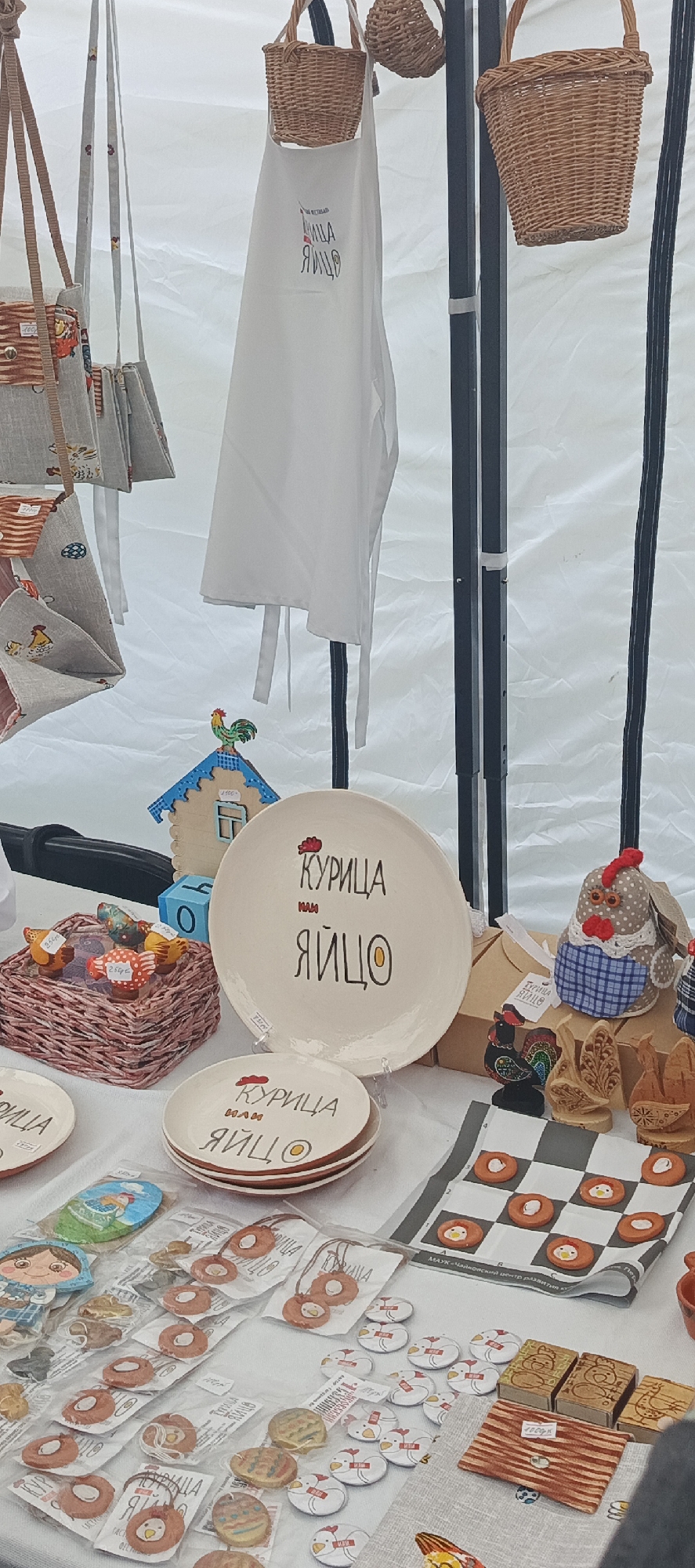 The image displays an unoccupied market stall exhibiting various handcrafted items, predominantly featuring a chicken and egg theme. The setting is indoors or under a canopy, indicated by a white, translucent fabric background and black metal support poles. In the upper section, a white apron, adorned with a red chicken head and the Cyrillic text "КУРИЦА или ЯЙЦО" (Chicken or Egg), hangs from a small woven basket. Another small woven basket hangs adjacent to it. To the left, several fabric bags or pouches with straps are suspended, some featuring striped patterns or small chicken/house motifs. One pouch has a price tag displaying "100 руб". The central display area features a large, cream-colored ceramic plate standing upright on a clear plastic easel. It bears the same "КУРИЦА или ЯЙЦО" design with a chicken head and an egg yolk, and a partial price tag indicates "1500 руб". In front of it, a stack of three smaller plates carries the identical design. To the left of these plates, a miniature wooden house with a blue roof and a colorful rooster figurine on top is visible. A price tag on the house reads "1500". Beside the house, a small woven basket contains several egg-shaped items, some with price tags of "250 руб" and "200 руб". On the right side of the central display, a fabric chicken doll with a red comb and blue plaid attire stands on a wooden block, featuring an attached tag. Next to it are several carved wooden items, including stylized roosters and other decorative pieces. A small wooden block with a painted rooster is also present. The foreground of the image showcases numerous smaller items arranged on a light-colored surface, possibly a tablecloth. These include: * A small fabric wallet with brown and white stripes, secured with a button, priced at "100 руб". * Several small wooden blocks or boxes with engraved chicken designs. * A checkered fabric placemat displaying nine small, round, orange-colored items with central chicken or egg designs, arranged in a grid pattern resembling a checkers board. * Numerous round white badges or pins, each depicting a red-combed chicken head, some also displaying "КУРИЦА или ЯЙЦО" in Cyrillic. * A selection of small items, individually sealed in clear plastic packaging. These include round pendants or keychains with chicken designs (labeled "КУРИЦА ИЛИ ЯЙЦО"), oval-shaped items with painted scenes (e.g., a figure of a girl, a landscape), and