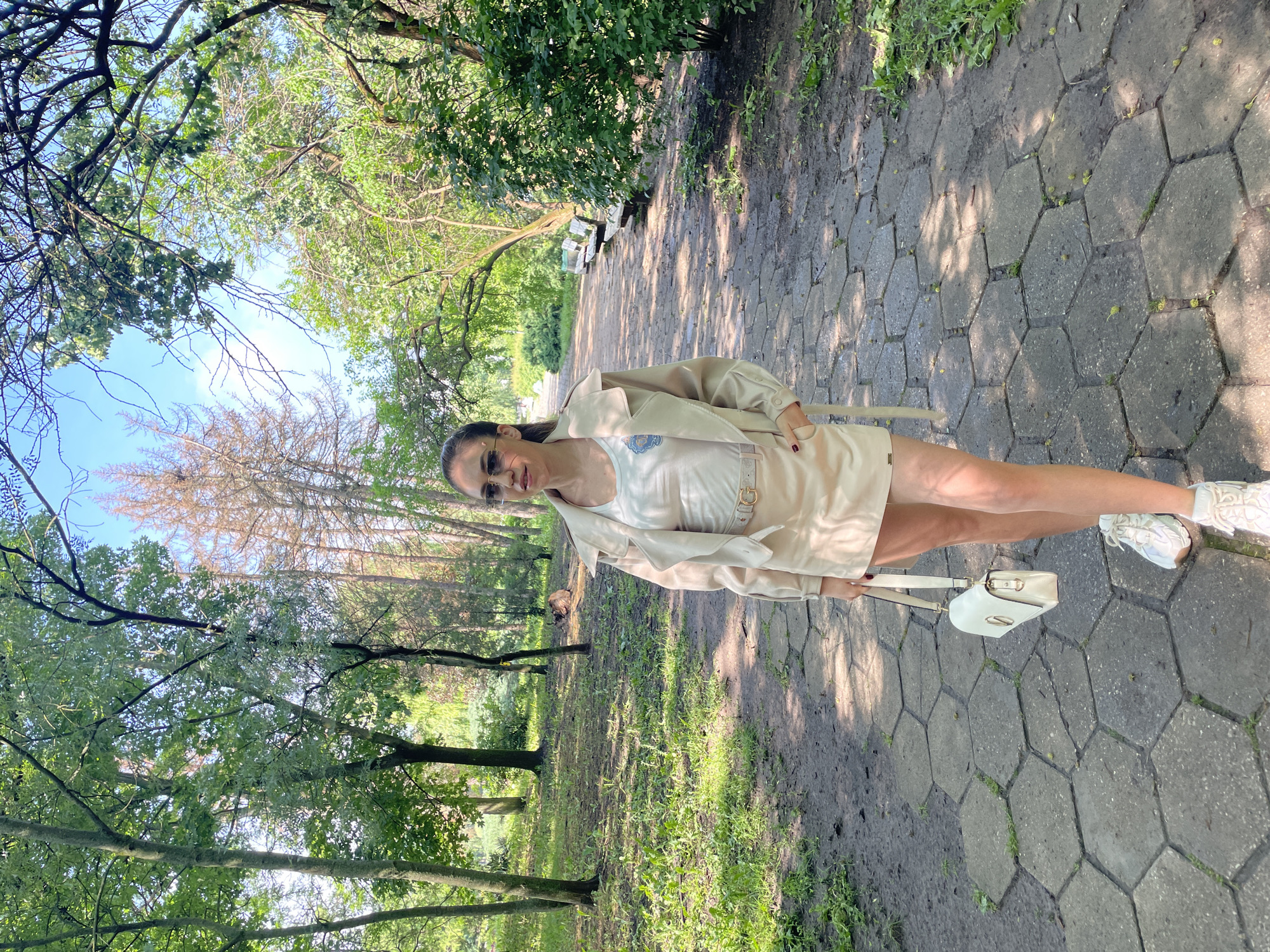 The image captures a woman standing on a paved path within a park in Sofia, Bulgaria, on a clear, sunny day. She is positioned in the foreground, facing the camera with a relaxed posture and a slight smile, accentuated by her sunglasses.
The woman has dark hair and is dressed in a light-colored ensemble, likely beige or cream, featuring a long-sleeved jacket, a white top adorned with a blue and white emblem, and matching shorts. She wears white athletic sneakers and carries a small, light-colored handbag with a distinct metallic closure. A belt with a prominent buckle is visible around her waist, and her fingernails are painted red.
The scene is an outdoor park environment. The path beneath her feet is paved with hexagonal concrete blocks, interspersed with small patches of grass and dirt. Lush green trees and foliage flank the path on both sides, creating a verdant setting. Glimpses of a bright blue sky are visible through the tree canopy, and the strong sunlight casts distinct shadows on the path, suggesting it is daytime, possibly mid-morning or late afternoon. Benches are visible further down the path, indicating a recreational area. The overall impression is one of a pleasant outdoor stroll in a natural setting. No discernible text is visible on the woman's clothing or accessories.