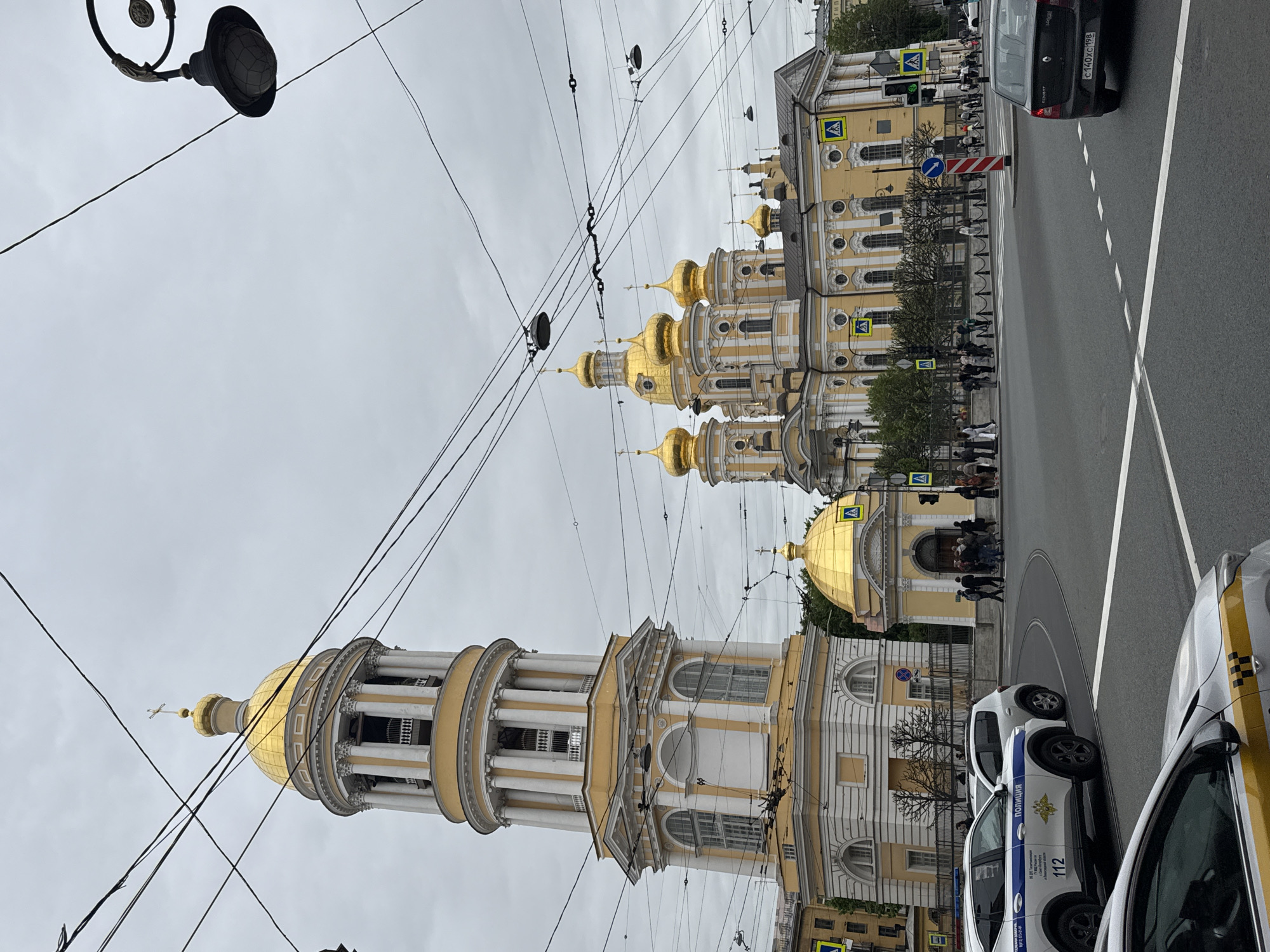 A street view in Saint Petersburg, Russia shows the Smolny Cathedral complex, its golden domes distinctive against a cloudy sky. Power lines crisscross the foreground. Pedestrians walk near the buildings. Several parked cars and a police vehicle are visible at the side of the road.