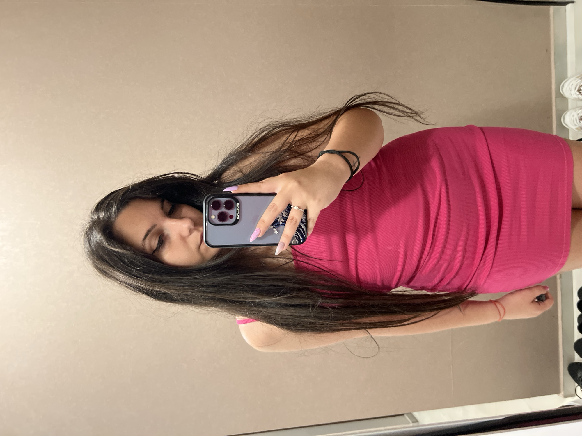 The image shows a young woman taking a selfie in a mirror. She has long, dark hair and is wearing a bright pink dress. She is holding a smartphone in her hand, and it is reflecting in the mirror. The background appears to be a room with light-colored walls. On the right side of the image, a rack of shoes is visible. The woman has long, light-colored fingernails and is wearing a ring on her left hand, and a thin red bracelet on her right wrist. The lighting suggests it is daytime. The location context is Sofia, Bulgaria.