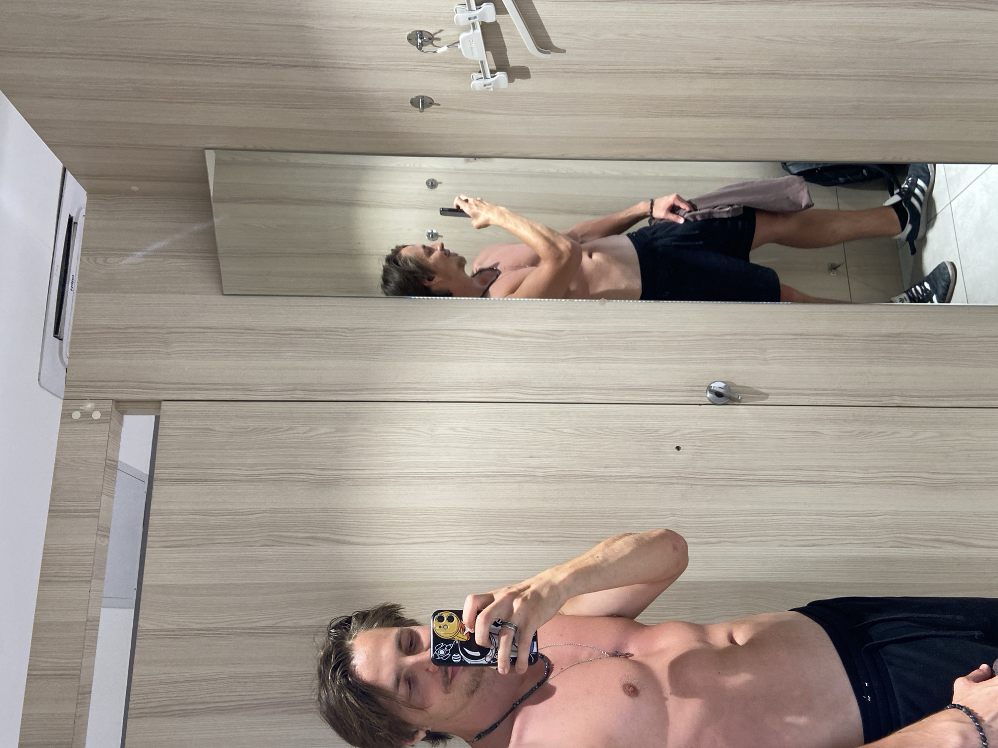A shirtless man in black shorts lies on his back, holding a smartphone and taking a selfie in front of a mirror inside a locker room. The locker room has light-colored wood-grain lockers. The location is Bjeliši, Montenegro.