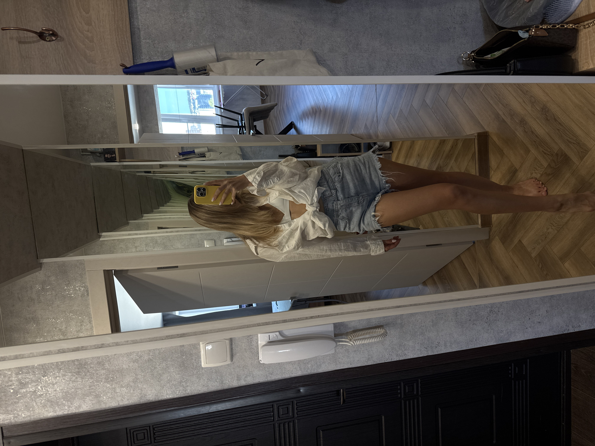A blonde woman in denim shorts and a white shirt is taking a picture of herself in a full-length mirror in what appears to be an apartment hallway. The hallway has wood flooring laid in a herringbone pattern. A white landline phone is mounted on the wall. A dark wood doorframe is visible at the bottom of the image. A lint roller, a cloth item, and a small handbag sit atop the mirror frame. A window is visible in the reflection.
