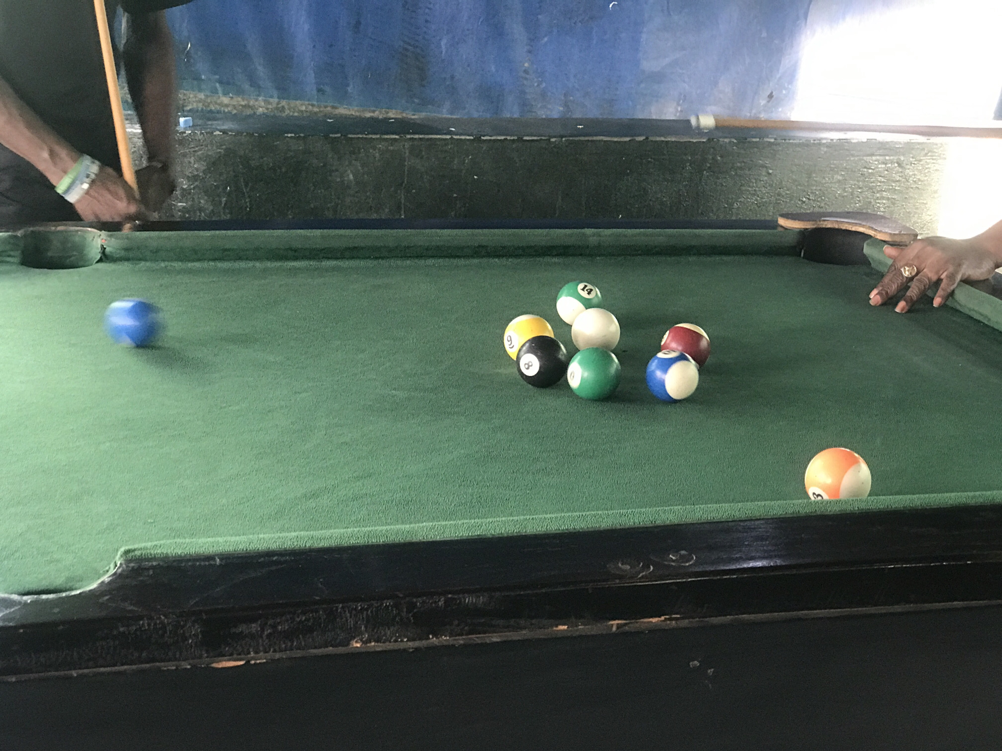 The image depicts a pool table with several billiard balls on its green felt surface. On the left side of the image, a person's arms and hands are visible, holding a pool cue and about to strike a blue ball. The person is wearing a wristband. In the center of the table, a cluster of balls is arranged, including a black 8-ball, a yellow 9-ball, a green 14-ball, a white ball, a red and white striped ball, a green ball, and a blue and white striped ball. To the right of these, a ball with an orange and white pattern, marked with the number 3, rests near the edge of the table. The background shows a blue wall with some textured details and what appears to be a white board or surface. The overall setting suggests a recreational area, possibly a bar or a game room in New Stadium, Nigeria.